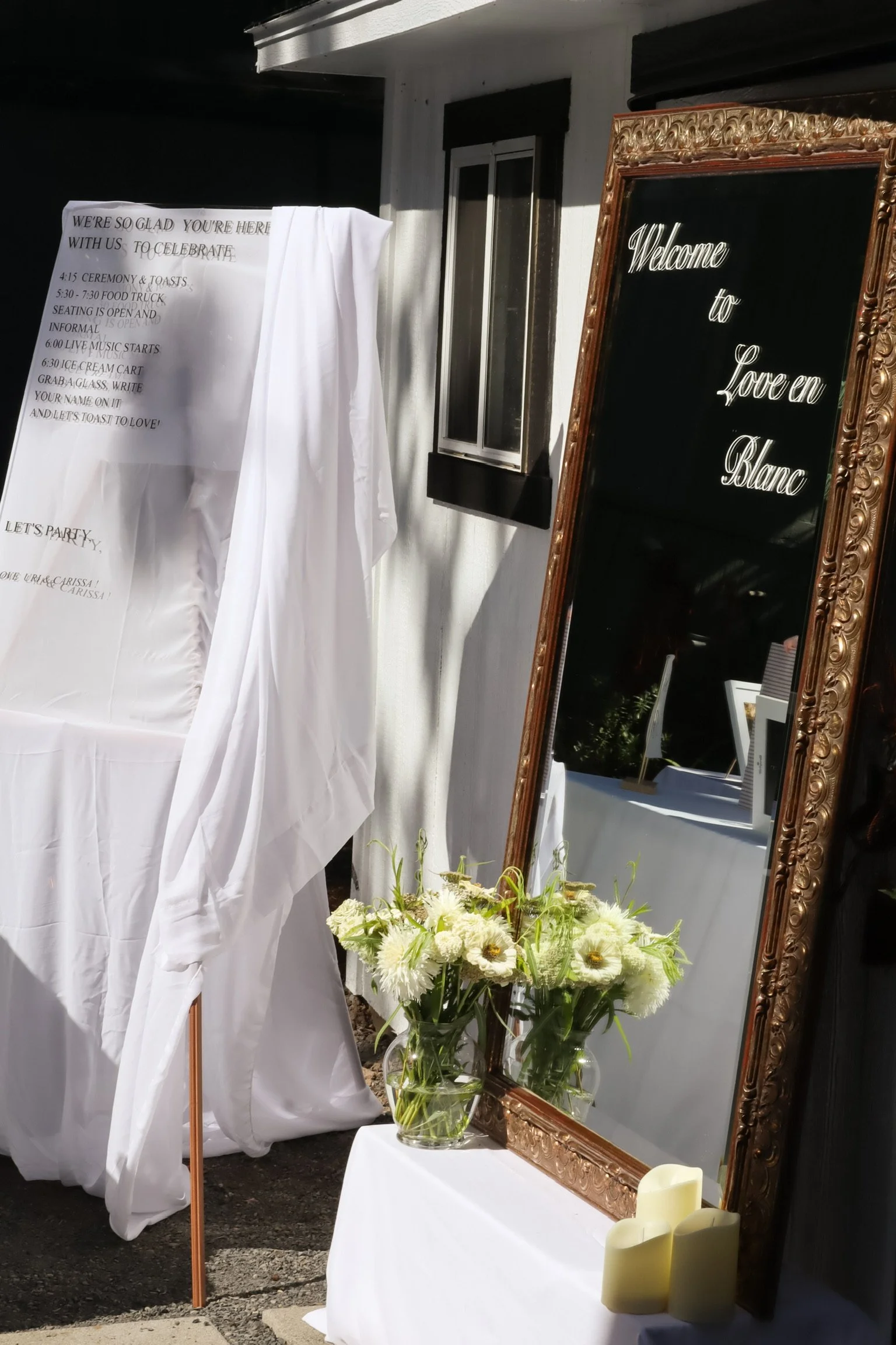 Decorative outdoor wedding setup with a large ornate mirror that has a welcome message, white floral arrangements in glass vases, candles, and a draped white tablecloth on a table, with a small window and black and white wall details in the backgroun