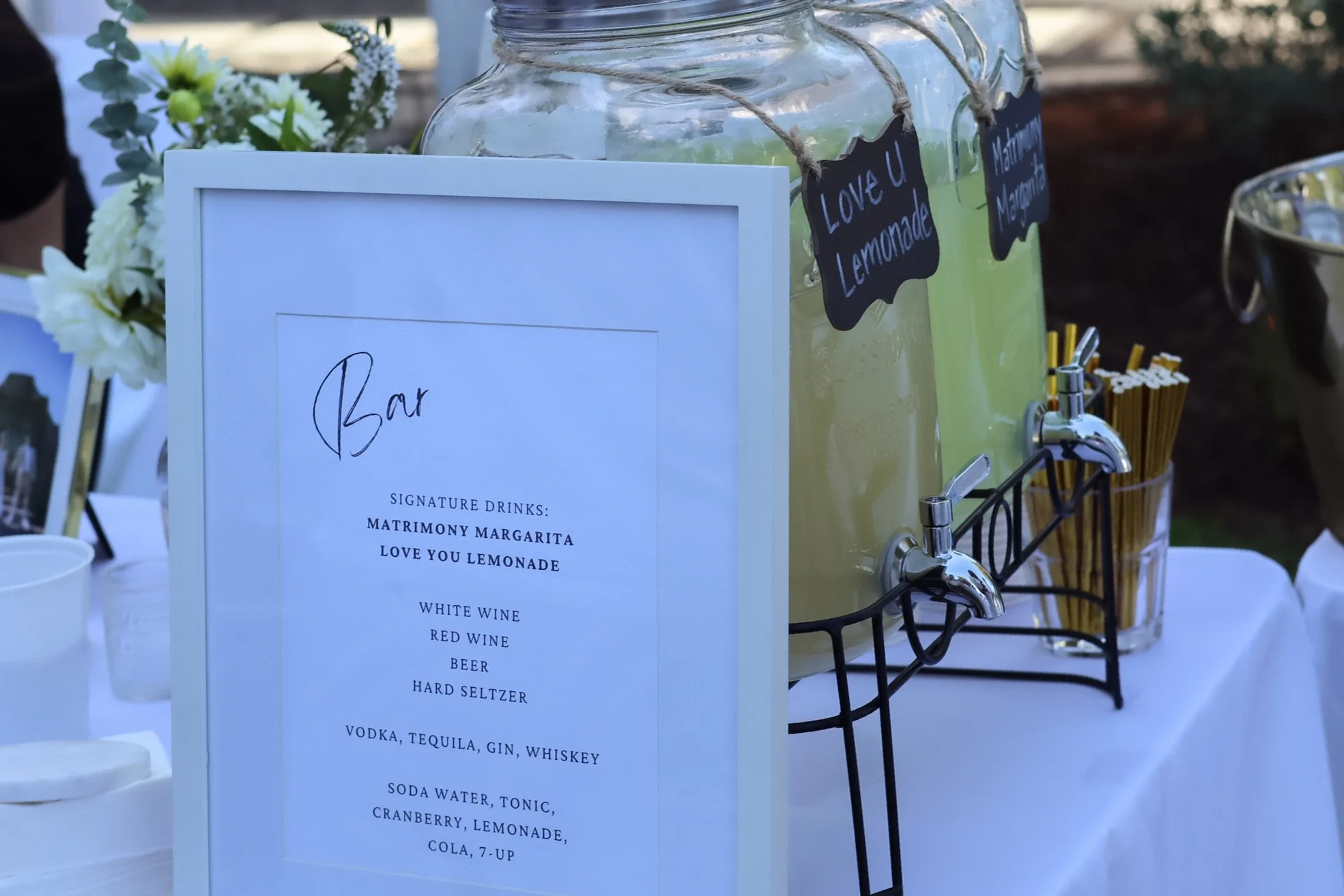 A drinks station with large glass jars containing lemonade, labeled 'Love U Lemonade' and 'Matrimony Margarita'. A white sign lists signature drinks including margarita, lemonade, wines, beer, seltzer, and various spirits, positioned in front of the 