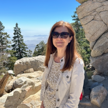 A woman with long gray hair, glasses, and a black outfit, standing against a gradient gray background.