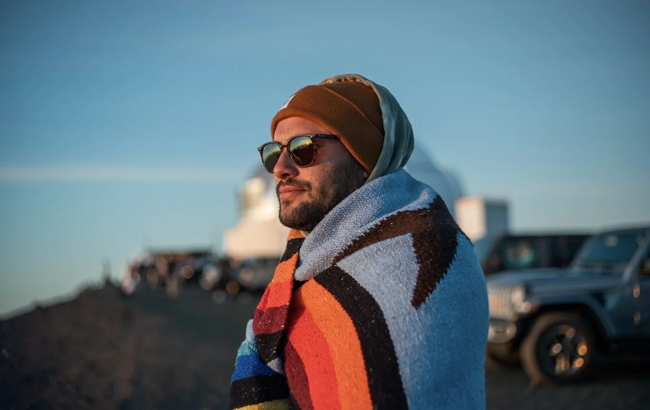 A man wearing sunglasses, a beanie, and a colorful striped blanket standing outdoors during sunset.