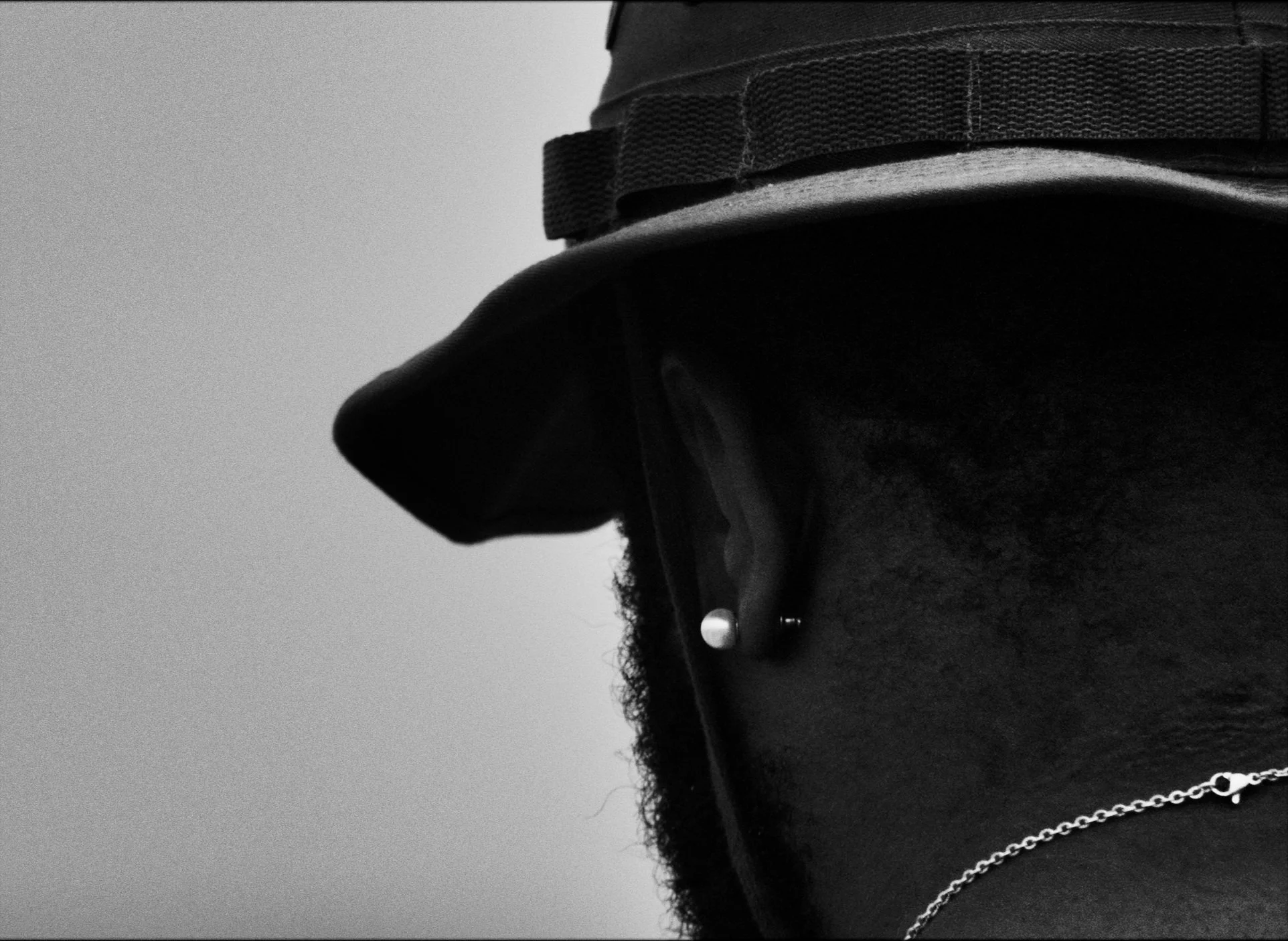 Close-up black-and-white photo of a person wearing a cap, with a chain necklace visible. The person's face is not shown.