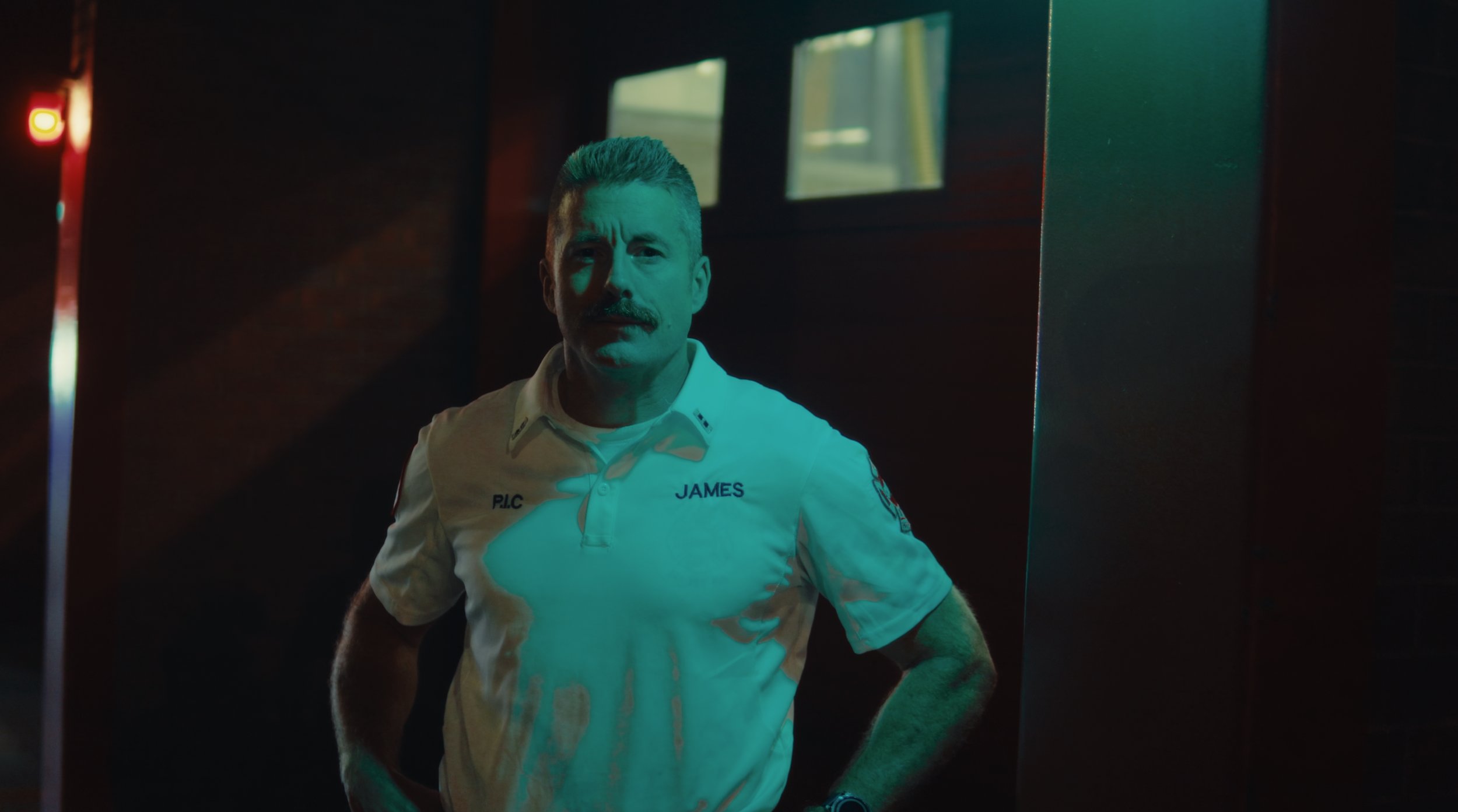 A man wearing a firefighter uniform with the name 'James' on it, standing with his hands on his hips in a dimly lit space with dark walls, illuminated by a bluish-green light.