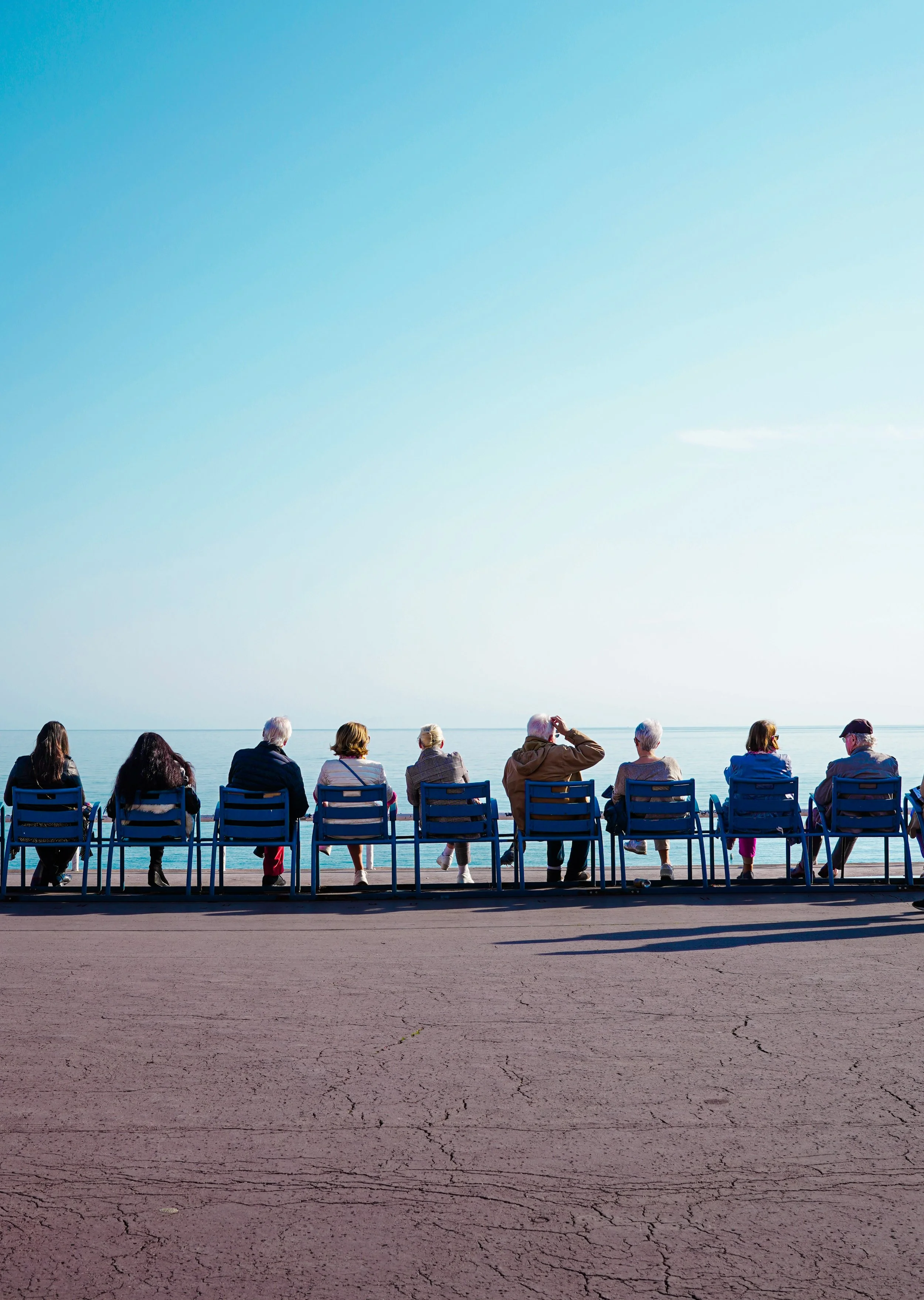 A group of people sitting on chairs facing a calm body of water, possibly at a seaside promenade on a clear day.