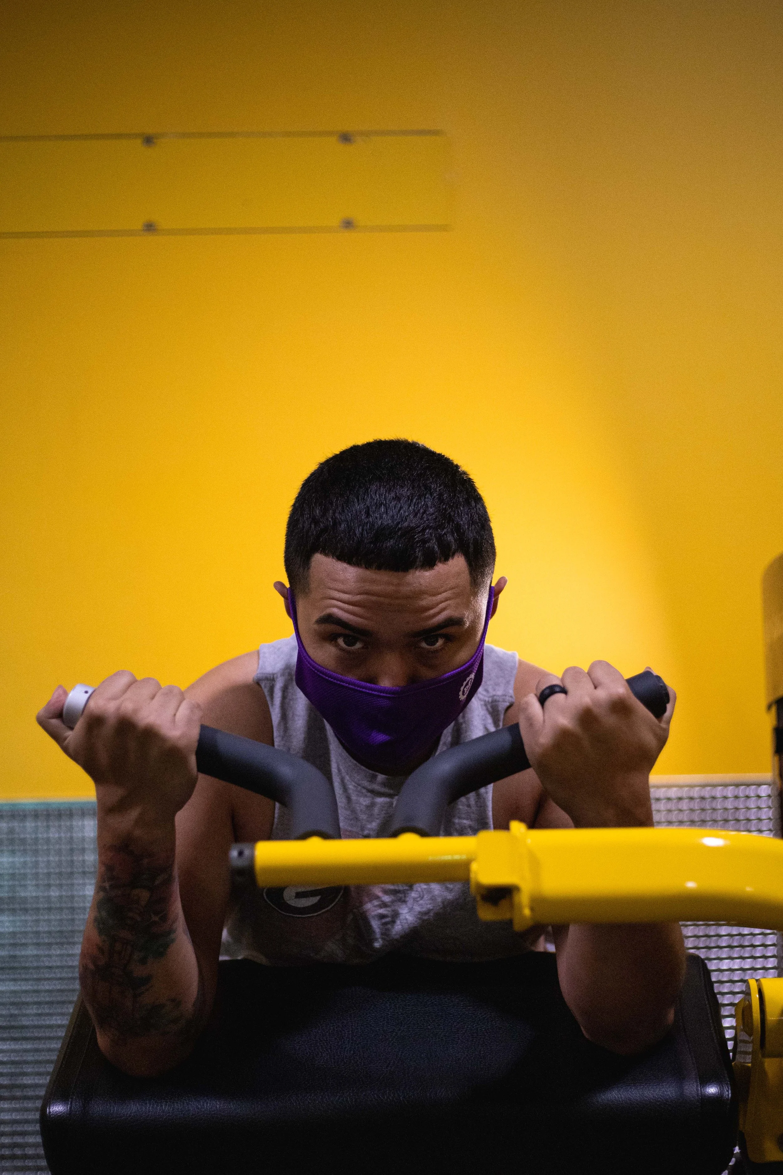 A man lifting weights at a gym with a bright yellow wall background, wearing a purple face mask and a sleeveless shirt.