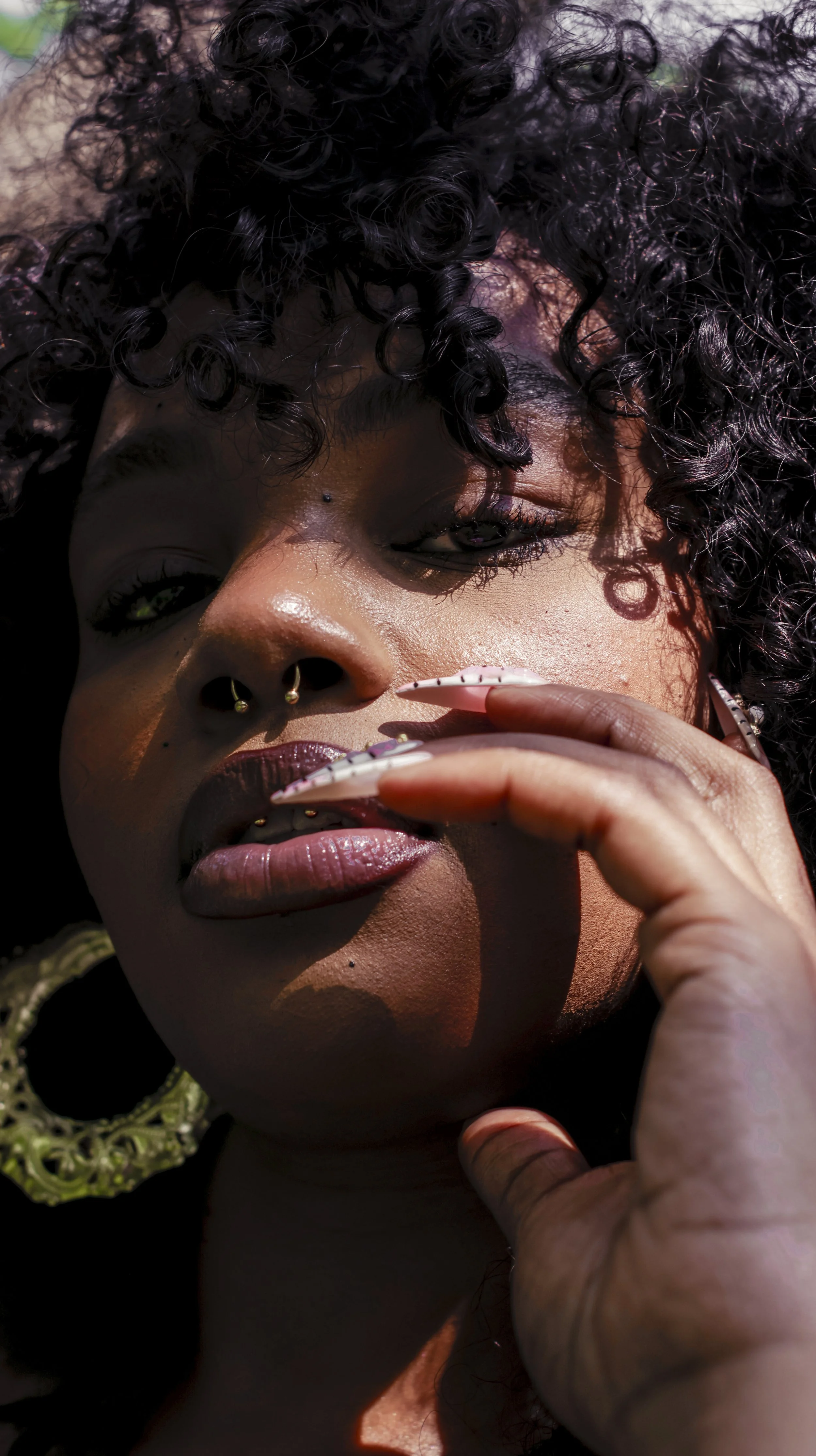 Close-up of a woman with curly hair, piercings, and artistic nails touching her lips.