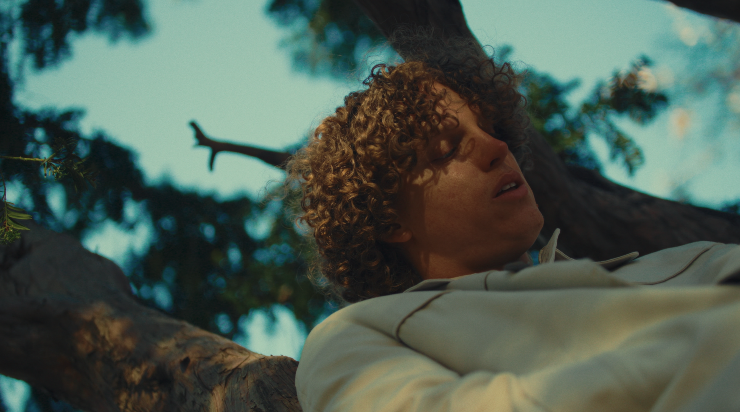 A person with curly hair leaning against a tree with their eyes closed, outdoors with a bright blue sky and leafy trees in the background.