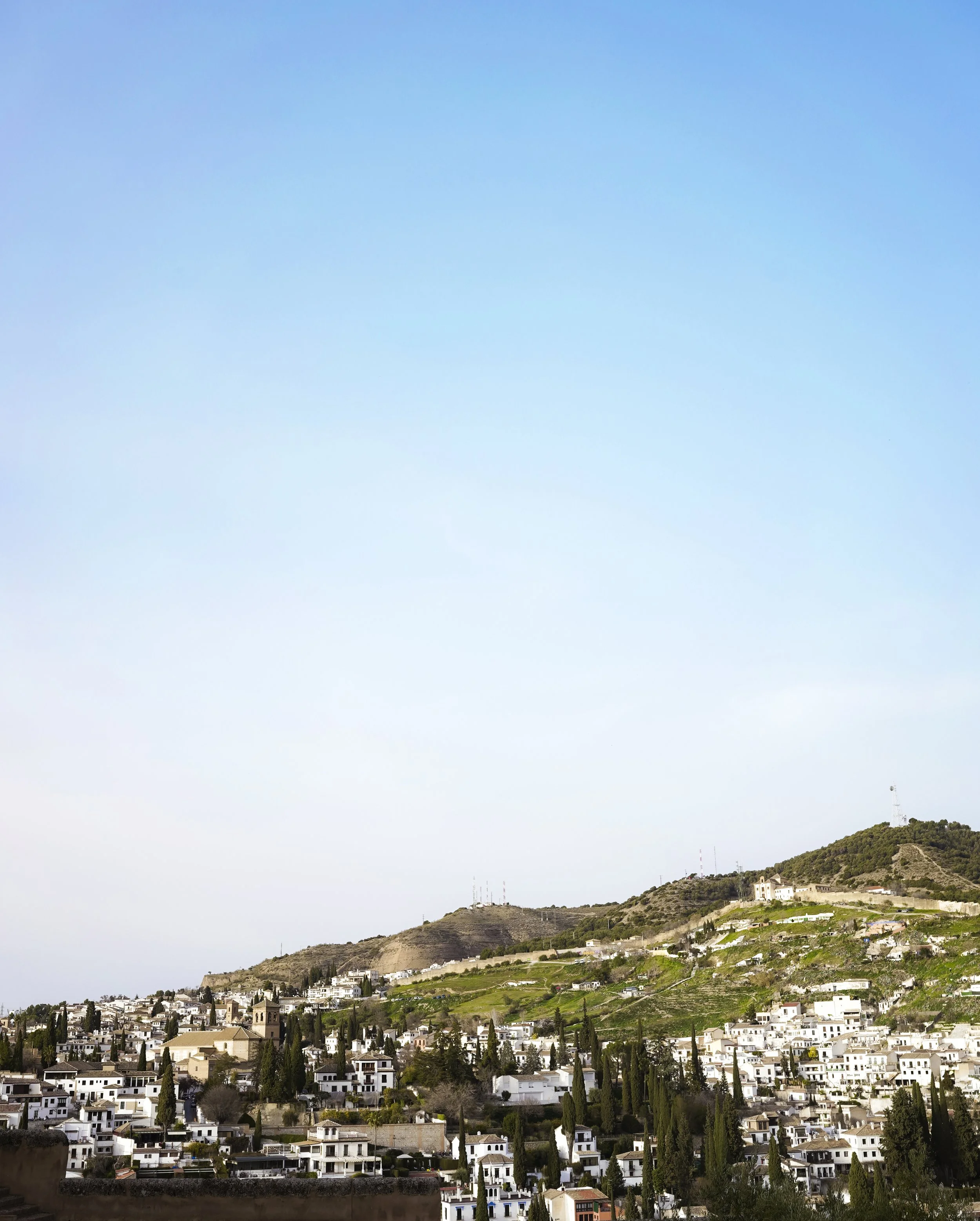 A scenic view of a hillside town with white buildings, green trees, and a mountain with communication towers in the background on a clear day.
