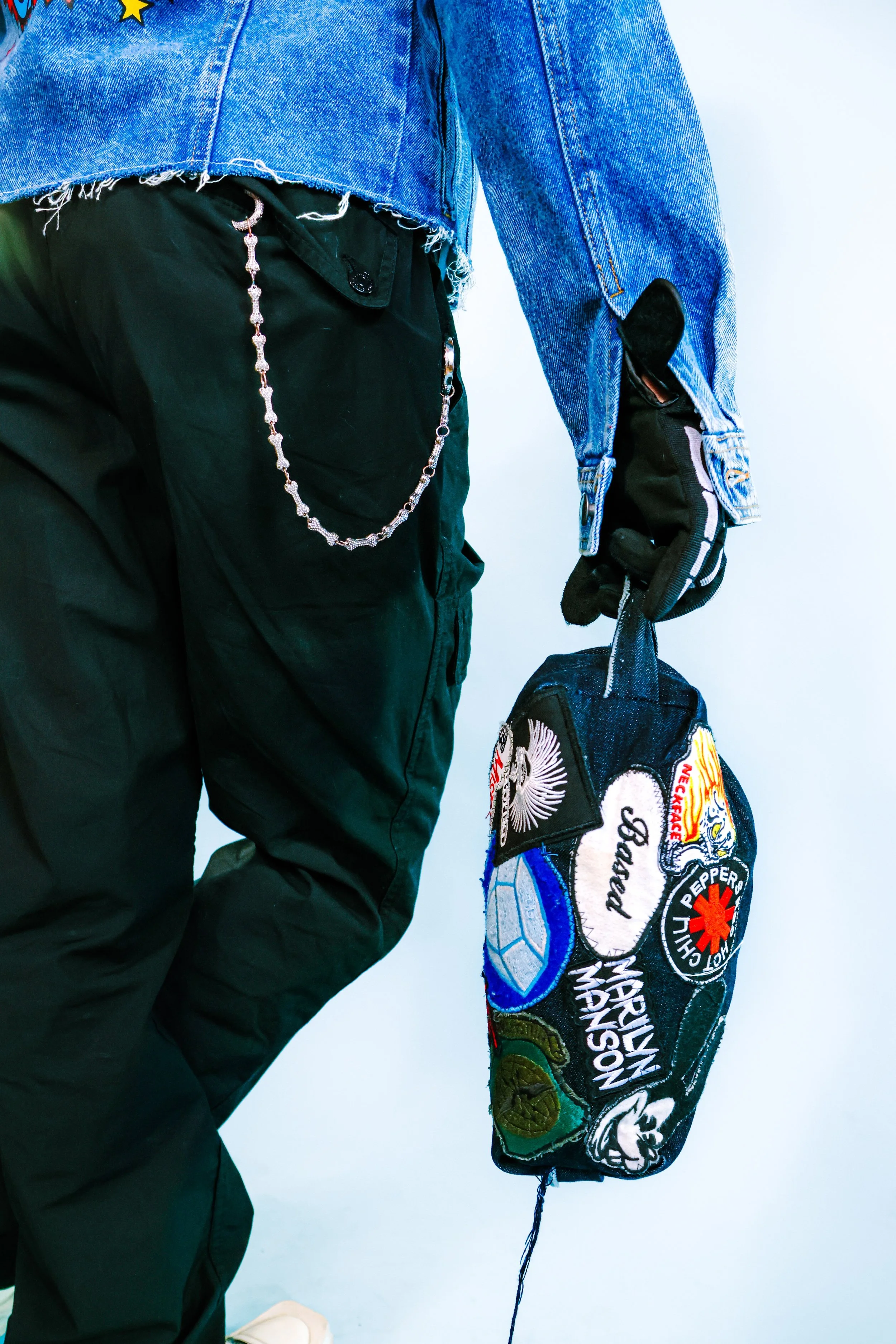 A person wearing a blue denim jacket, black pants with a silver chain, and gloves holding a denim bomber bag decorated with various patches.