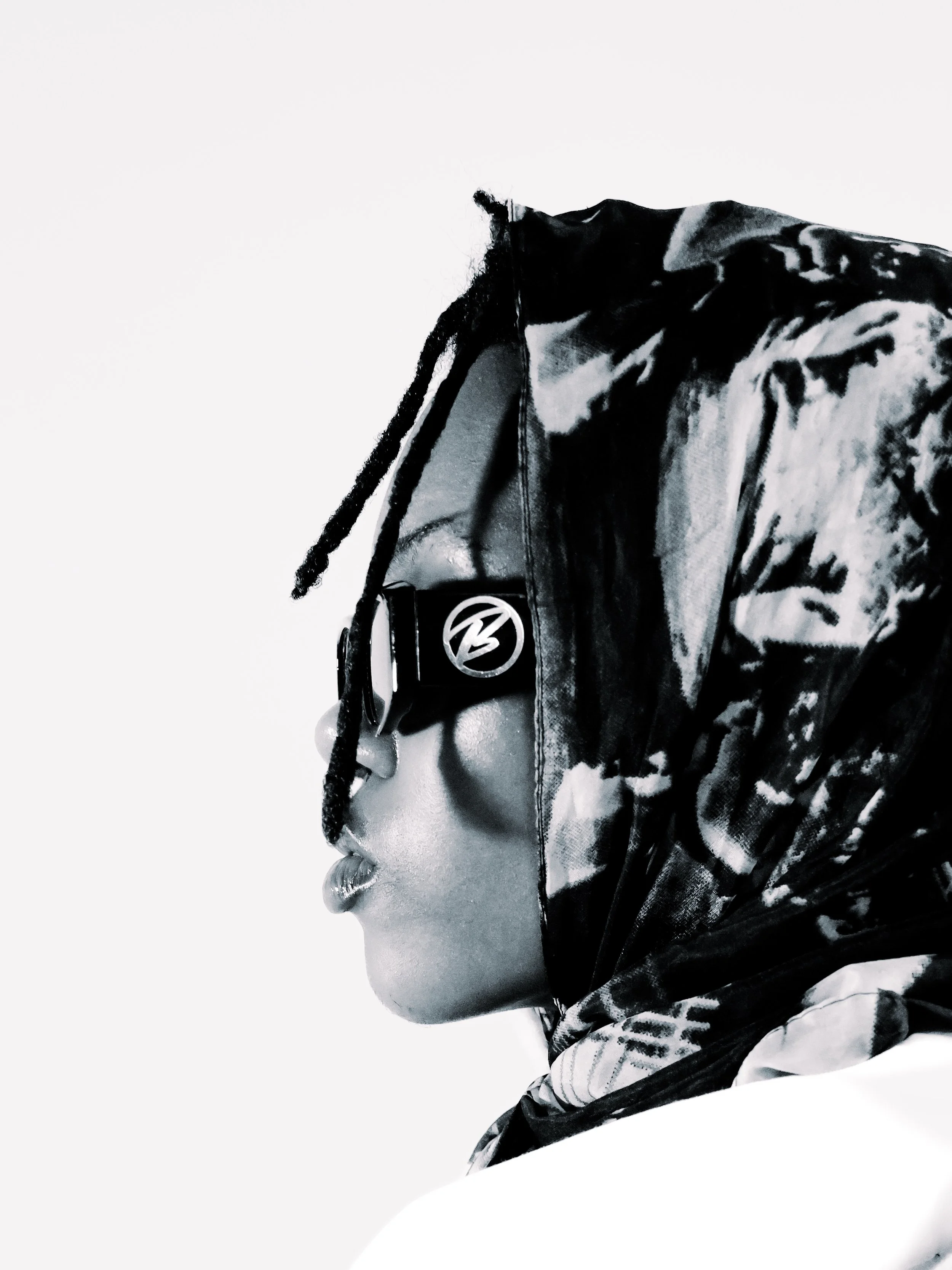 Side profile of a woman wearing sunglasses, a patterned headscarf, and a jacket, in black and white.