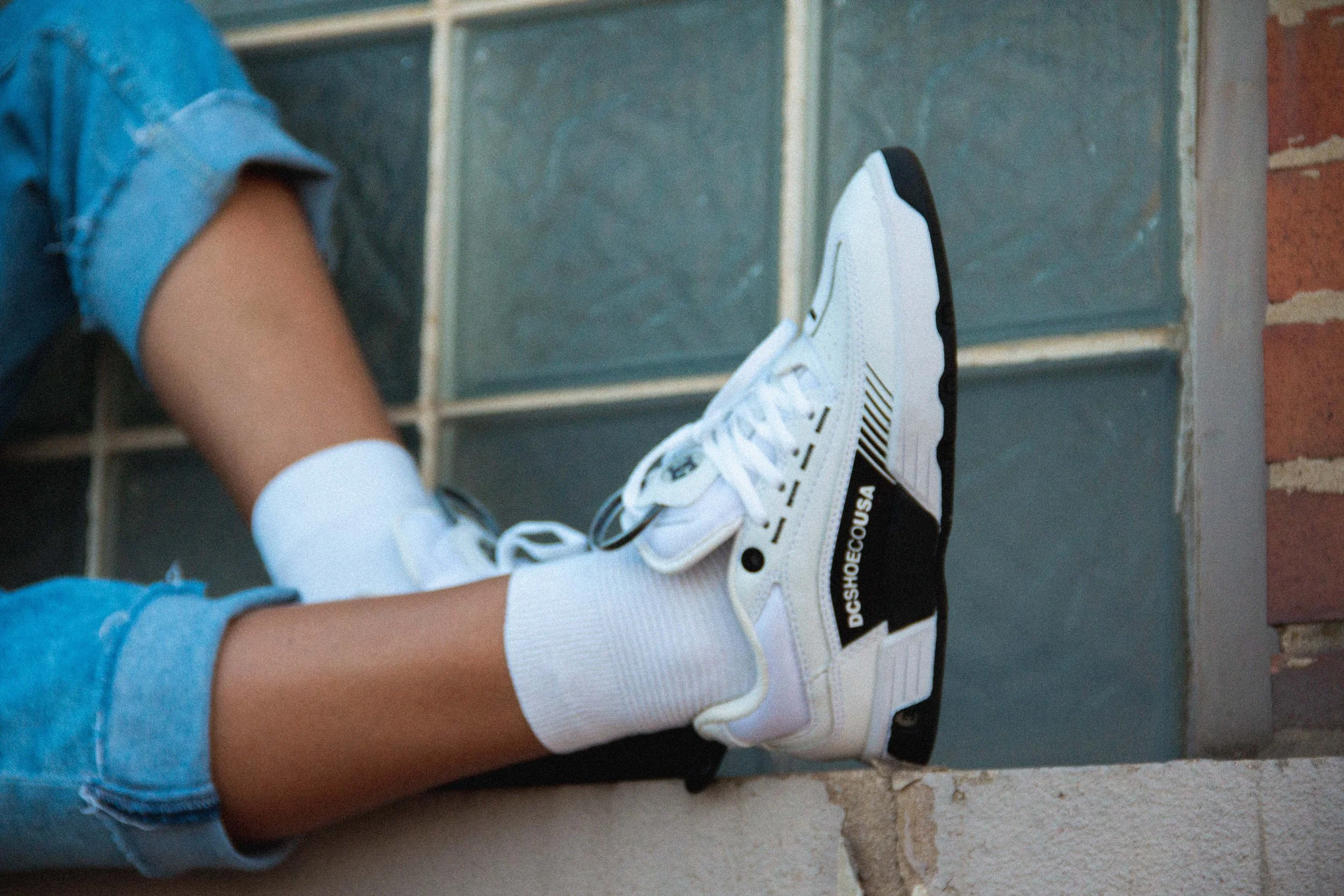 Close-up of a person's legs wearing distressed denim shorts and white socks, with white DC Shoes sneakers featuring black accents and the text "DCSHOECOUSA" on the side, sitting on a brick ledge in front of a glass block wall.