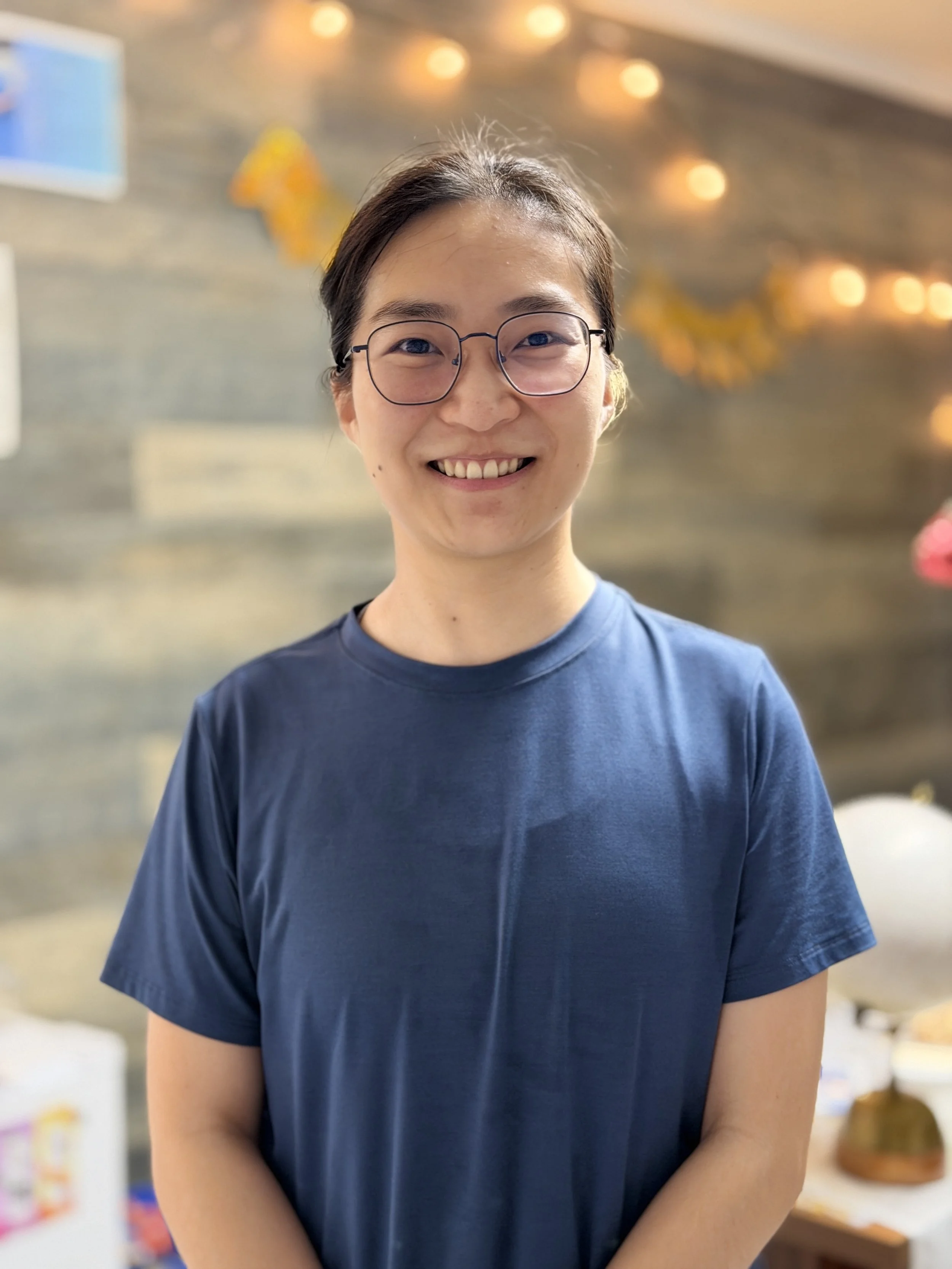 A smiling woman with glasses and dark hair tied back, wearing a blue t-shirt, standing in front of a background decorated with string lights and paper decorations.