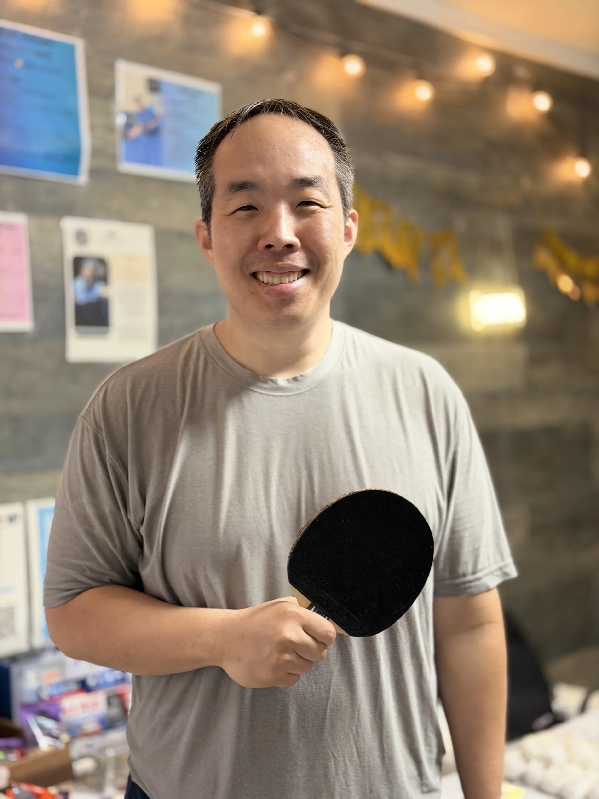 A man smiling and holding a paddle in a room decorated with string lights and posters on the wall.
