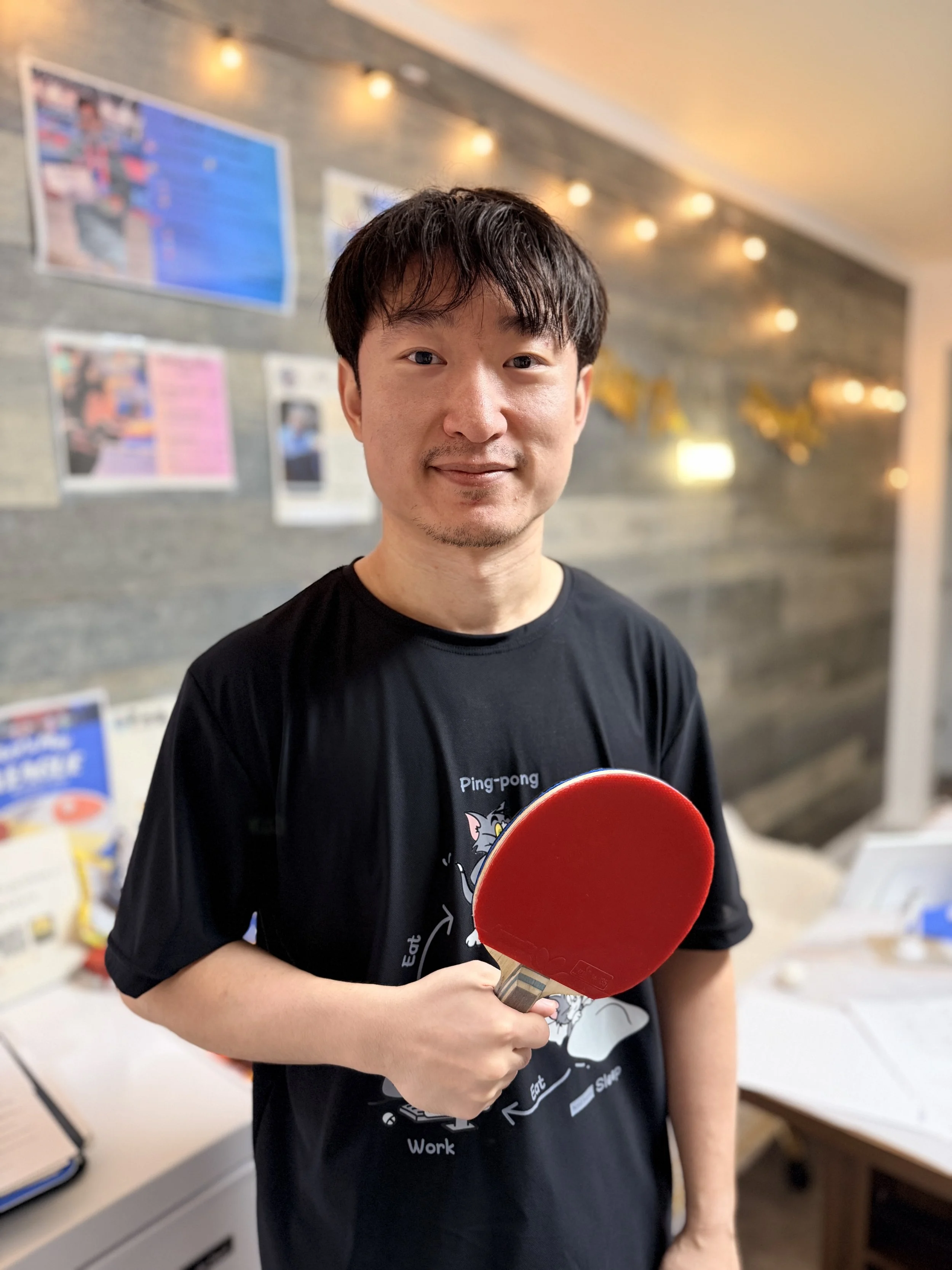 A man holding a red ping pong paddle inside a room with a gray brick wall and blurred posters in the background.