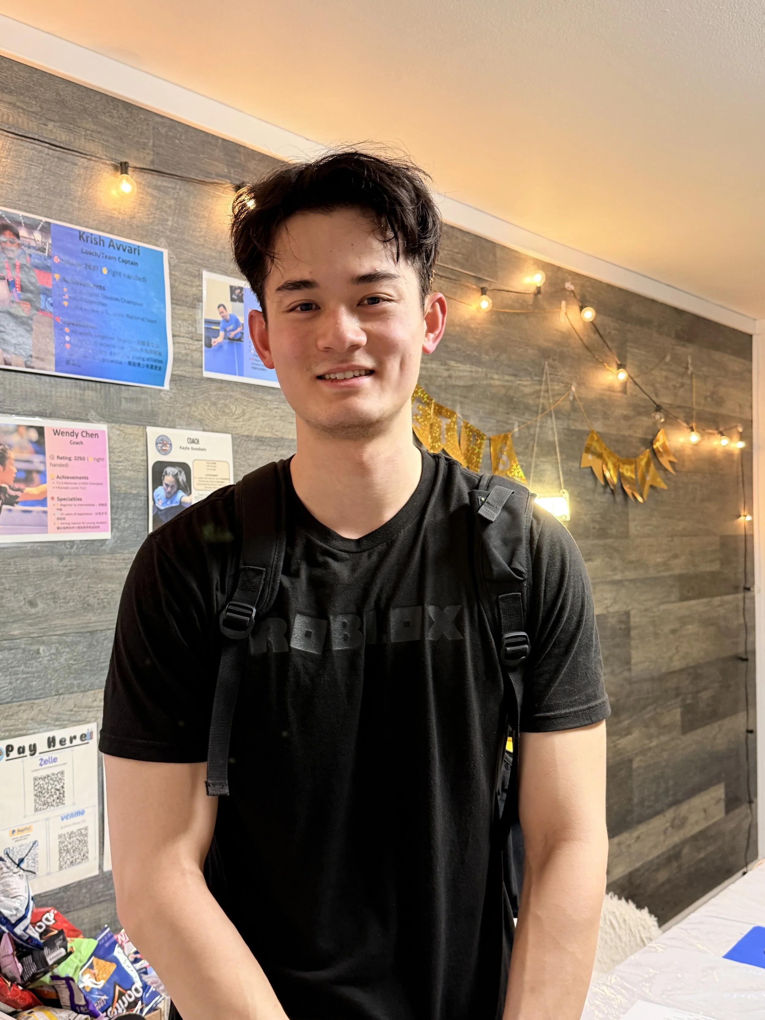 A young man smiling, wearing a black t-shirt and a backpack, standing indoors with a wood-paneled wall behind him decorated with string lights and a gold banner.