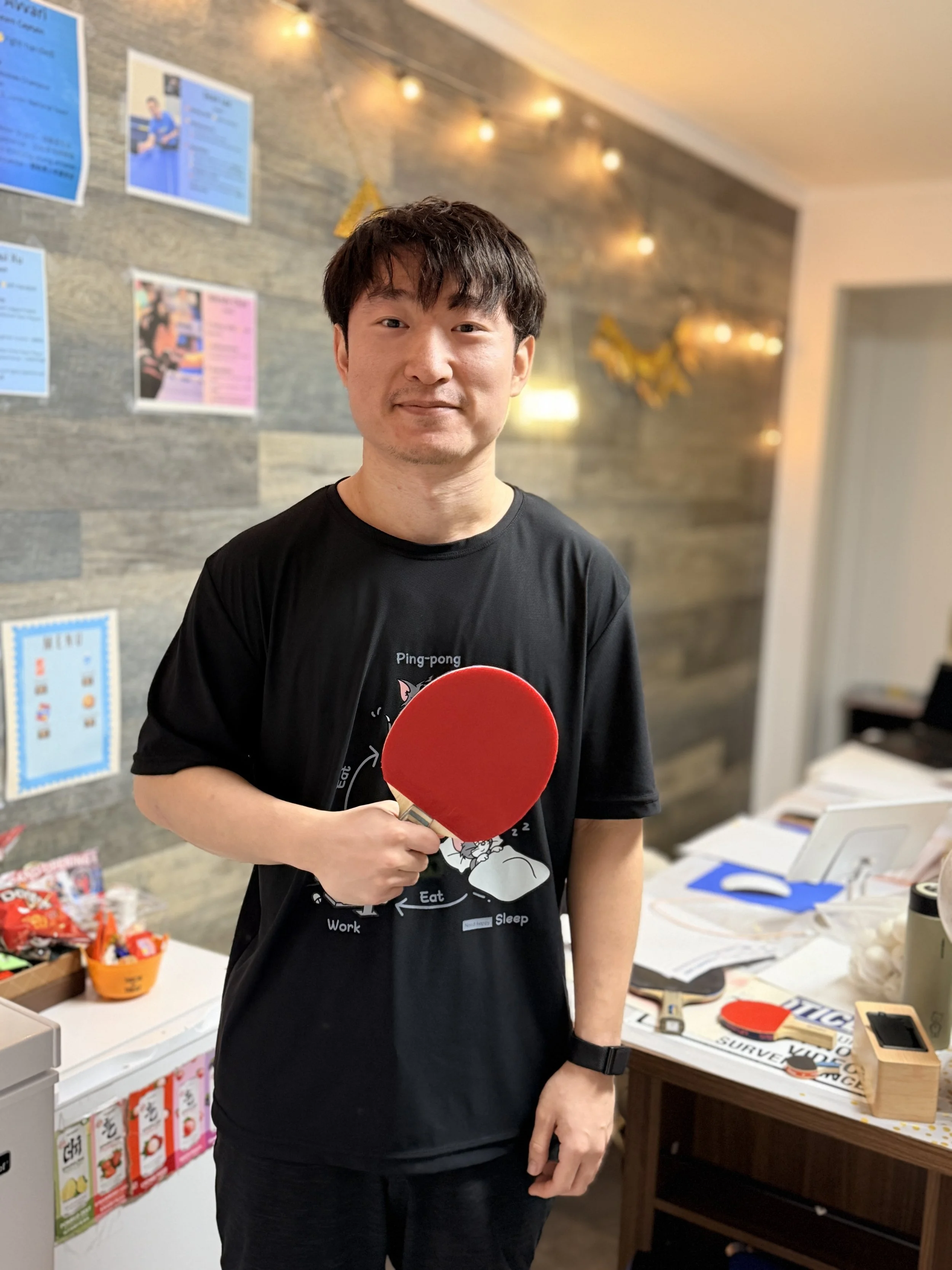 A man holding a red ping-pong paddle standing in an indoor setting with a wooden wall and decorations behind him.