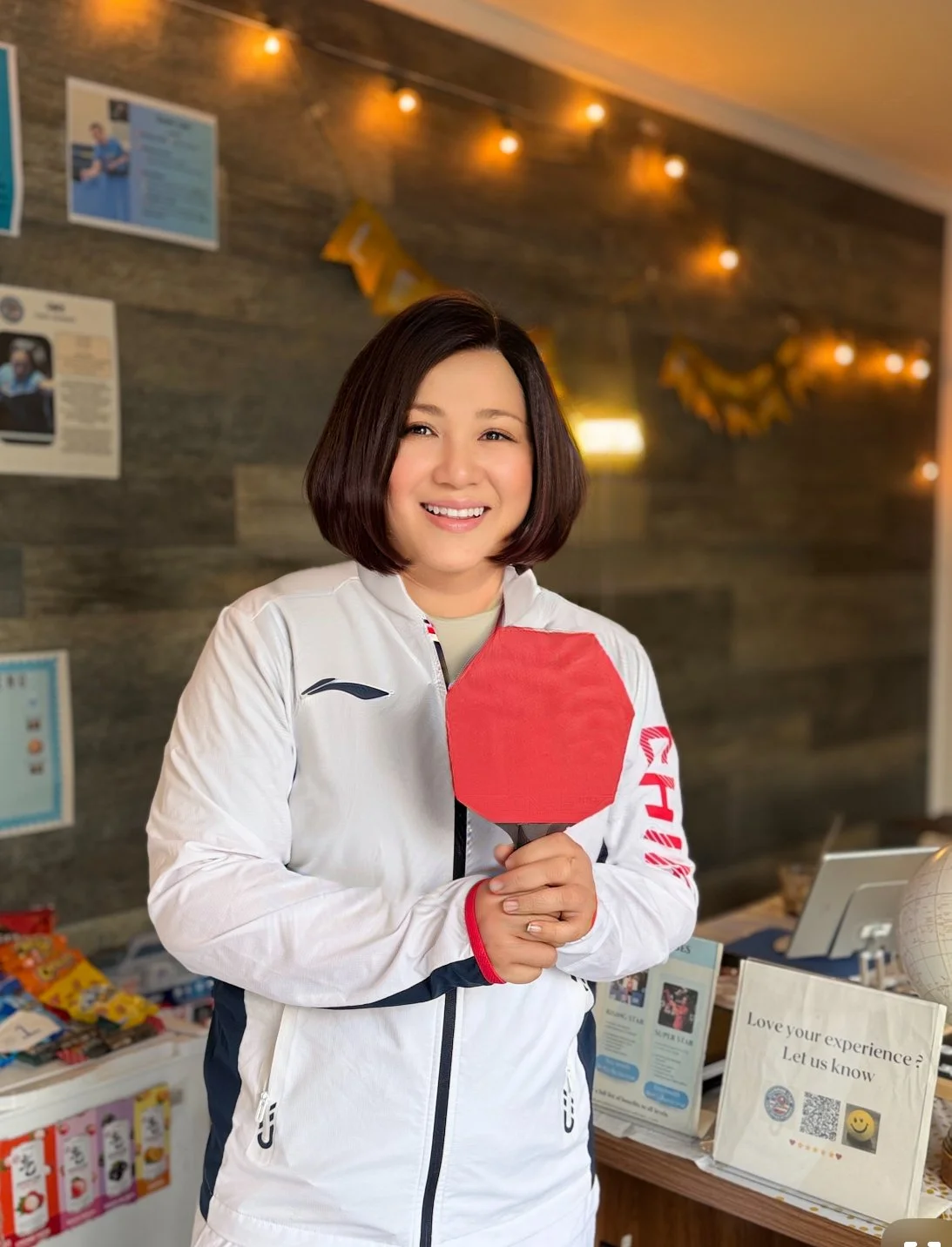 A woman smiling and holding a red table tennis paddle inside an indoor space decorated with string lights and posters.