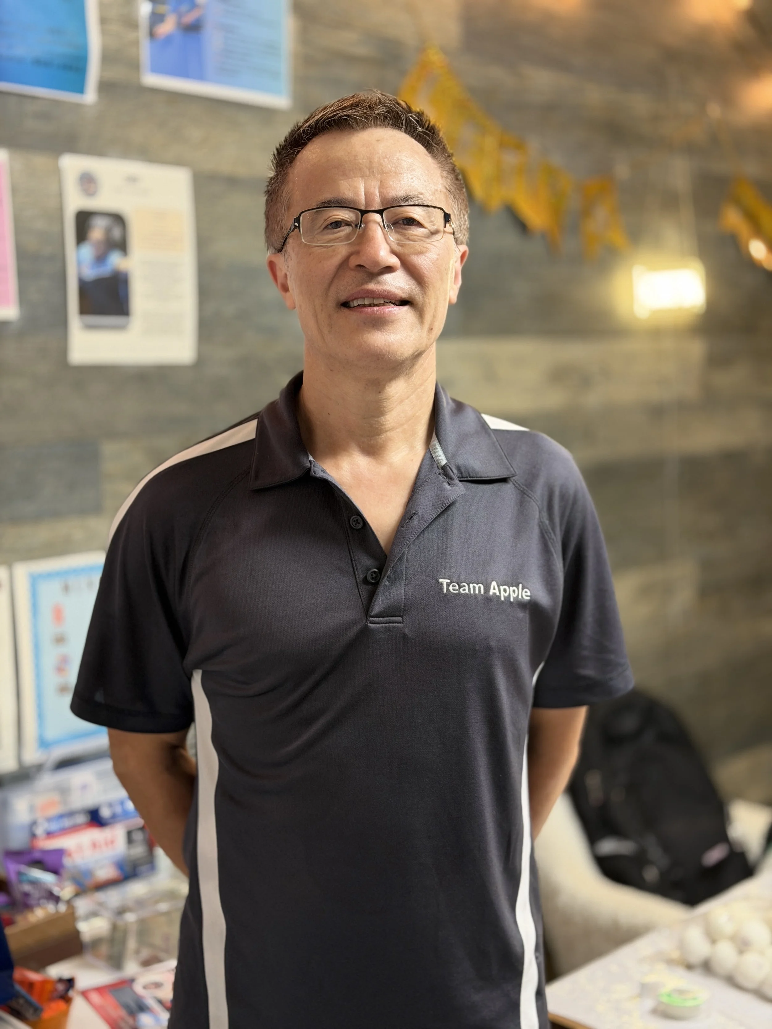 A man in glasses wearing a black polo shirt with white accents and 'Team Apple' embroidered on it, standing indoors in front of a wall with posters and decorations.