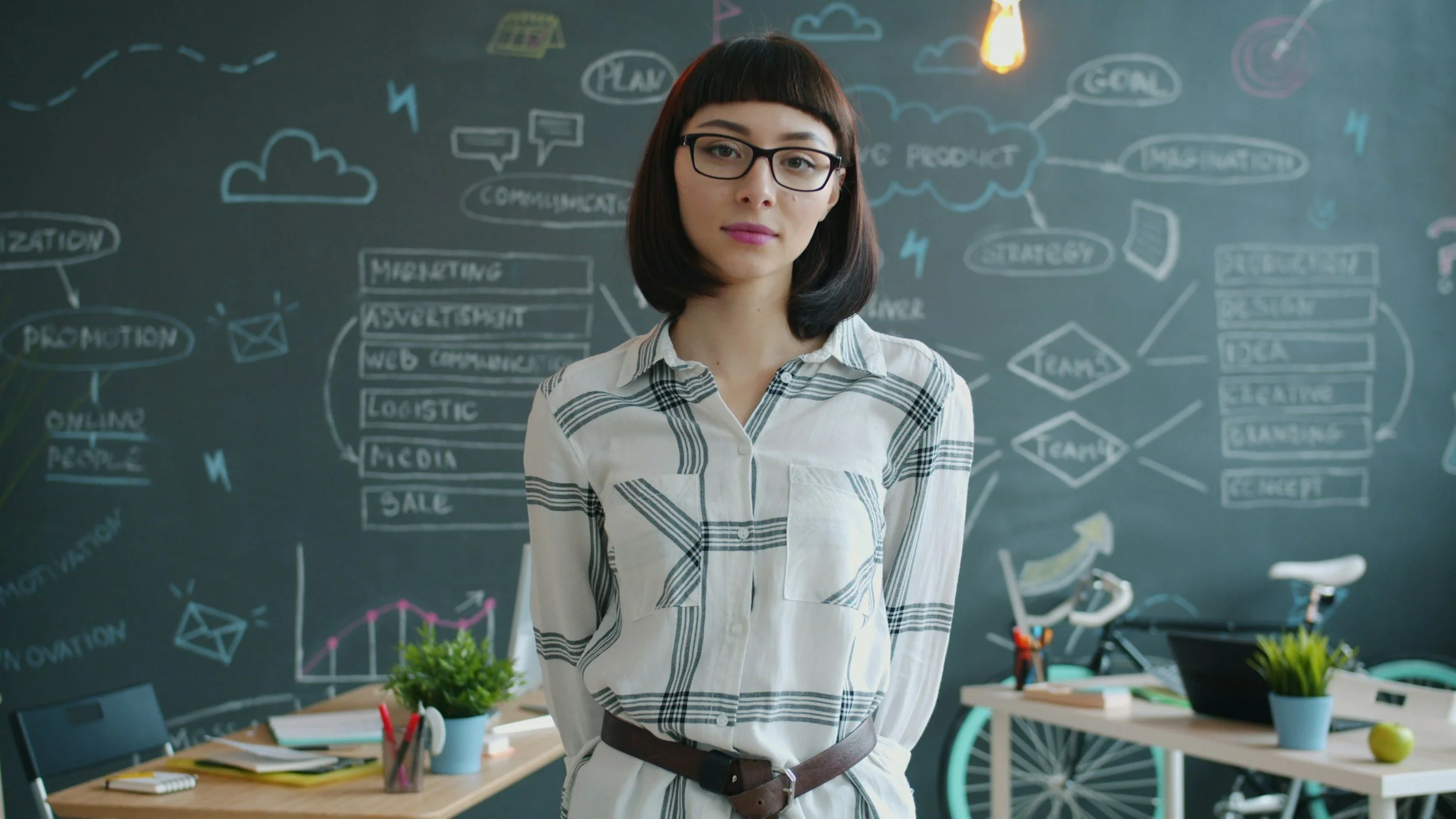 A woman wearing glasses stands in a room with a chalkboard background filled with business and marketing diagrams. The room has desks with plants, office supplies, and bicycles in the background.
