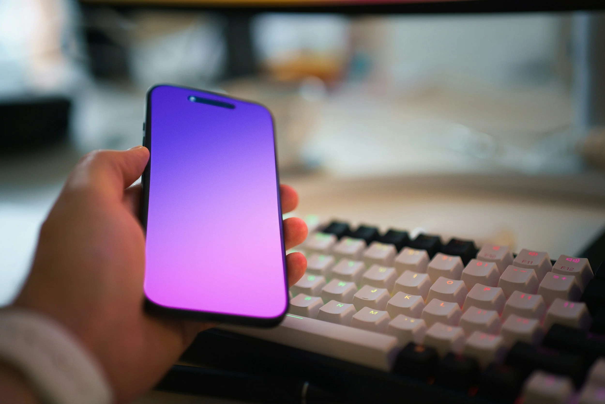 A hand holding a smartphone with a gradient purple and pink screen, in a workspace with a white mechanical keyboard with colorful backlit keys in the background.
