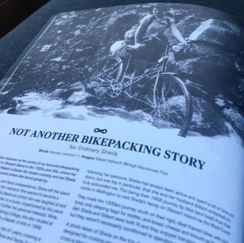 Photograph of a magazine page with a black and white image of a person riding a mountain bike through water, accompanied by a story titled 'Not Another Bikepacking Story'. Hannah is the author.