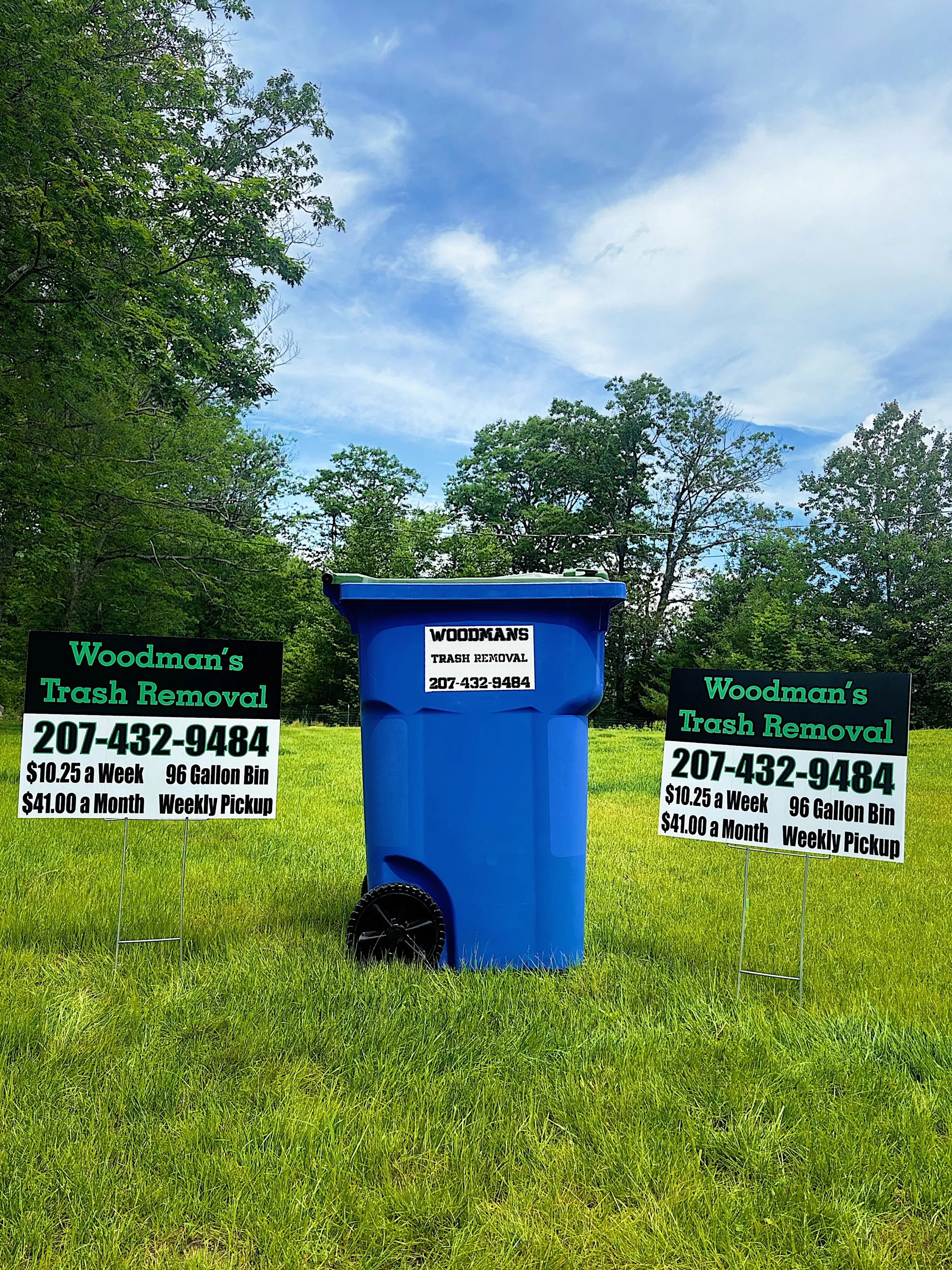 Blue trash bin with signs on either side advertising Woodman's Trash Removal with a phone number, price, and pickup options, set in a grassy field with trees and a cloudy sky in the background.