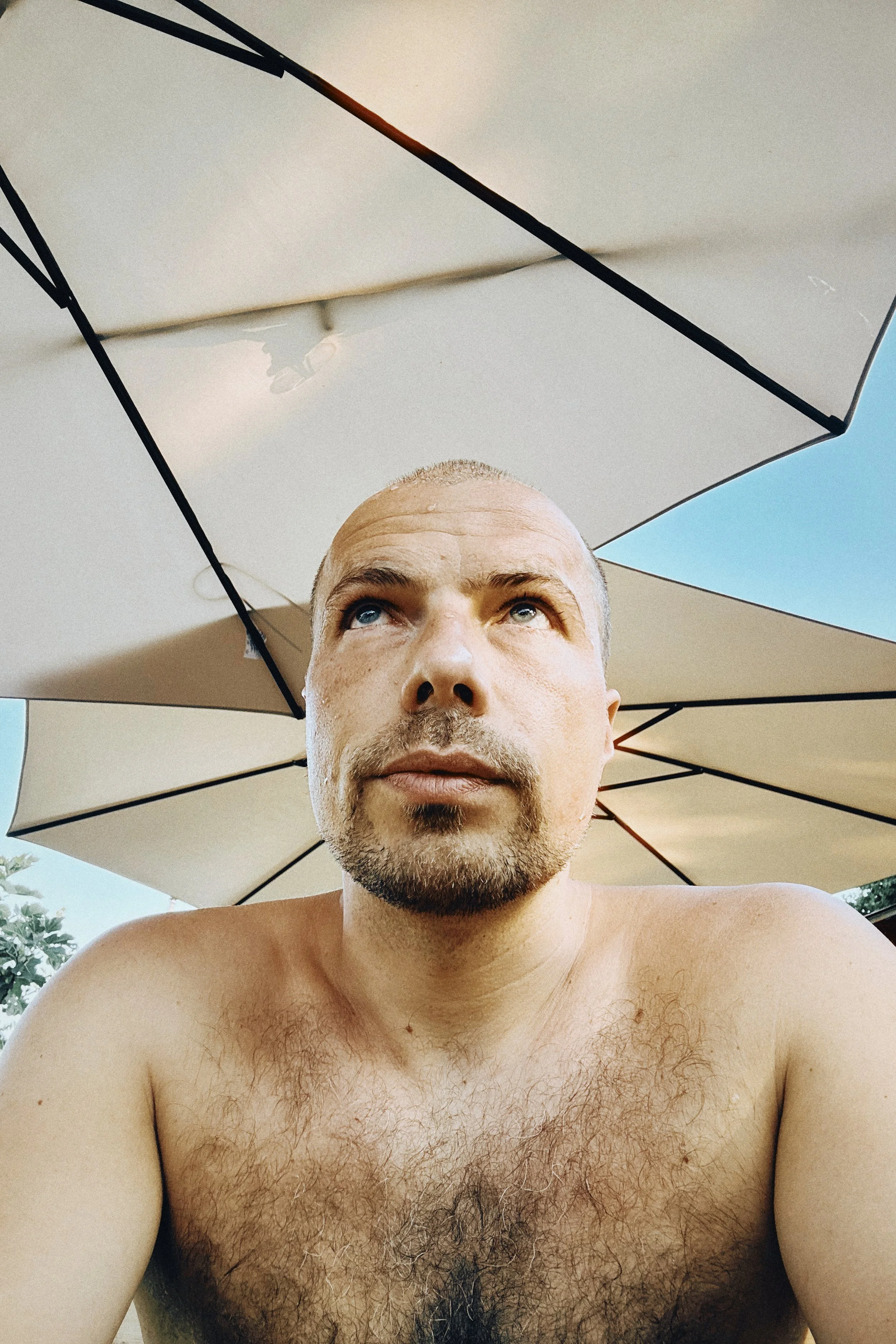 Nahaufnahme eines Mannes ohne Oberteil, mit kurz blondem Haar, Bart und sichtbarem Brusthaar, im Außenbereich unter einem großen Sonnenschirm, blauer Himmel im Hintergrund.