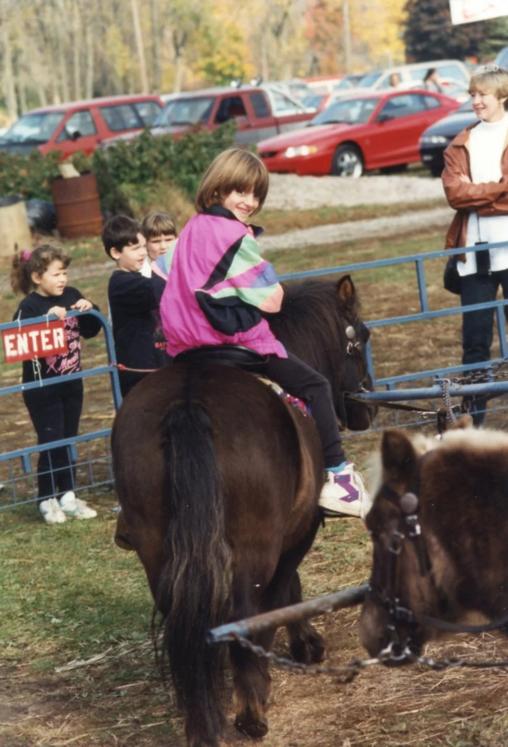 Me at 5ish years old on a live pony carousel ride at a fair. I turn to look over my shoulder at the camera from the saddle with a big smile on my face. I'm wearing a very 90s pink windbreaker