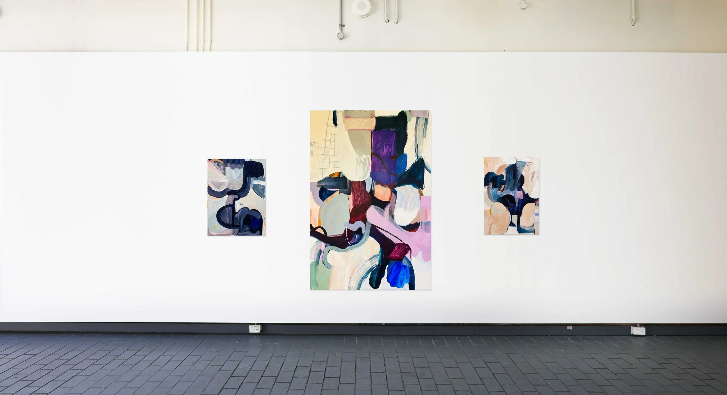 Gallery wall featuring three abstract paintings with vibrant colors, hung against a white wall in an art gallery space.