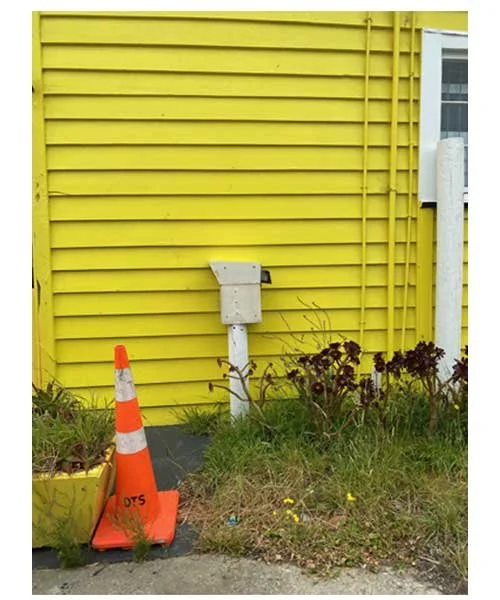 Yellow building with overhead wiring, an orange traffic cone, a yellow flowerpot, and some plants and weeds in the grass