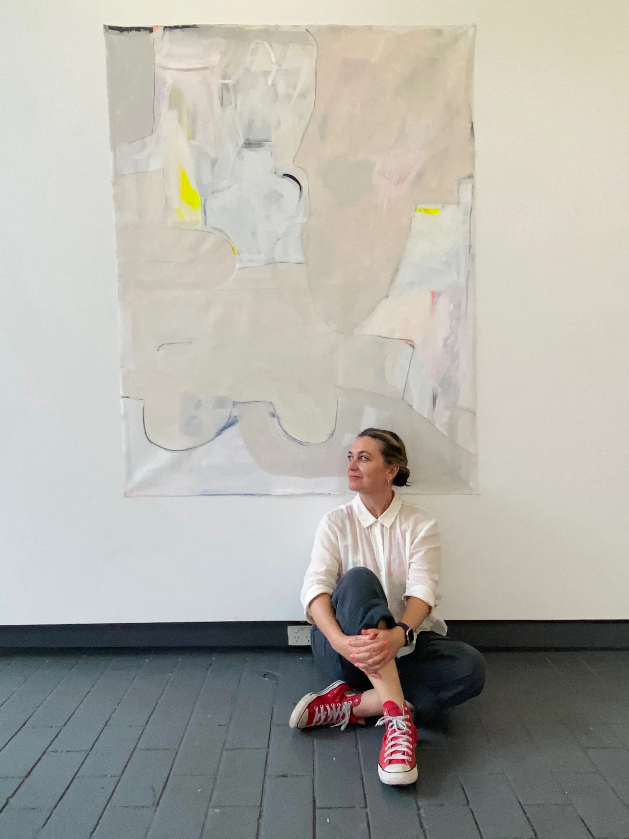 A woman with dark blonde hair tied back, wearing a white shirt, black pants, and red sneakers, sitting cross-legged on a dark wooden floor in front of an abstract painting on a white wall.