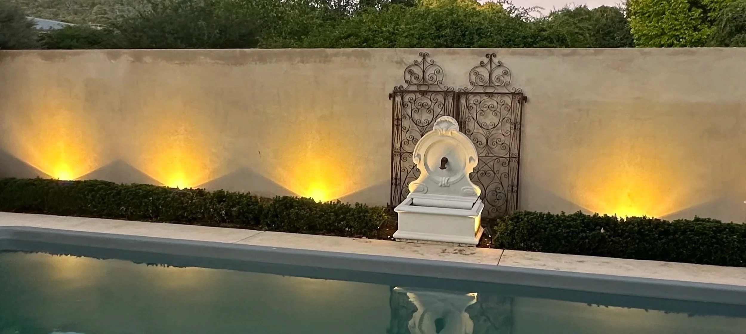 Night view of a backyard with a swimming pool, illuminated garden lights, and a decorative white fountain with an ornate metal gate behind it, all against a beige wall with greenery in the background.