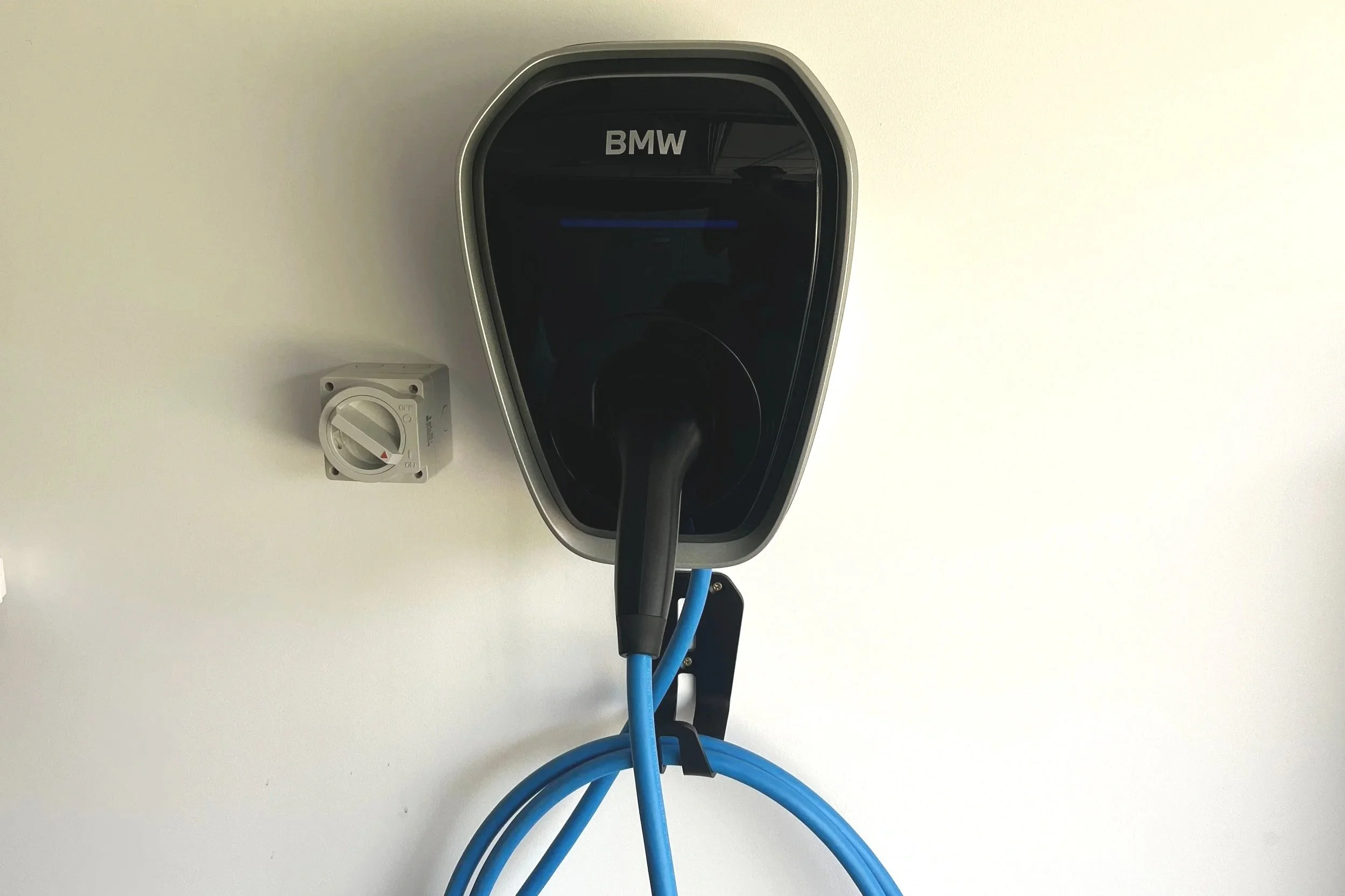 An electric vehicle charging station on a white wall with a black charging port labeled "BMW" and a blue charging cable connected.