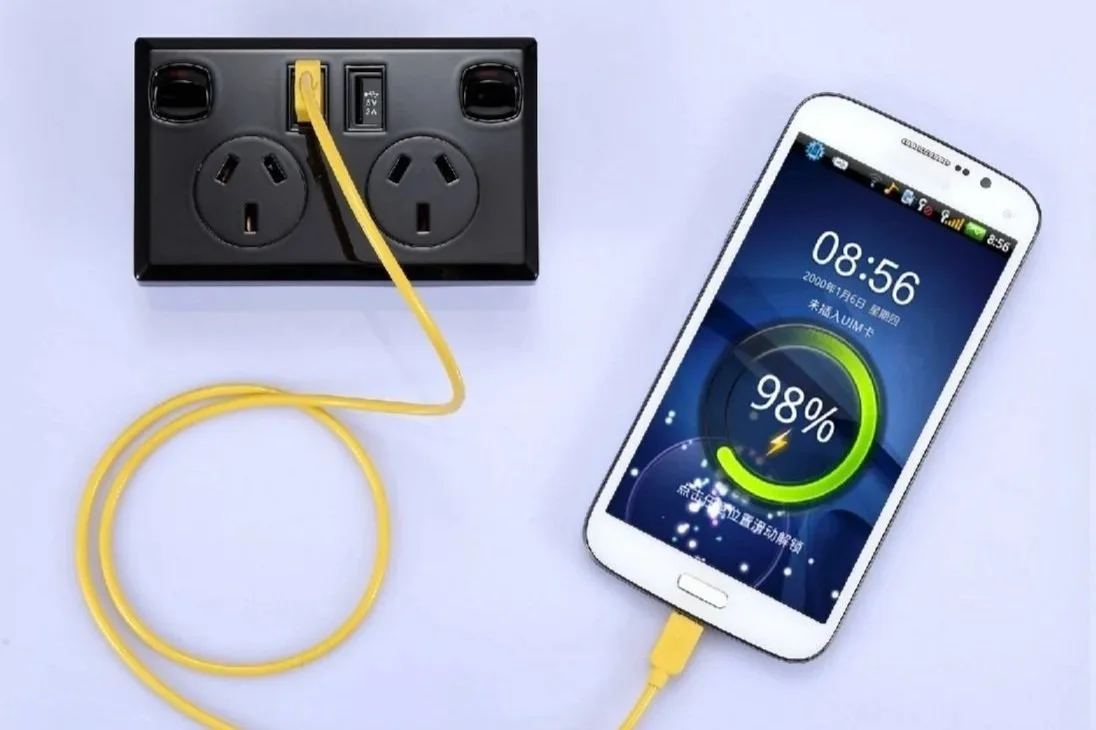 A smartphone charging through a yellow charging cable plugged into a white wall outlet with two sockets.