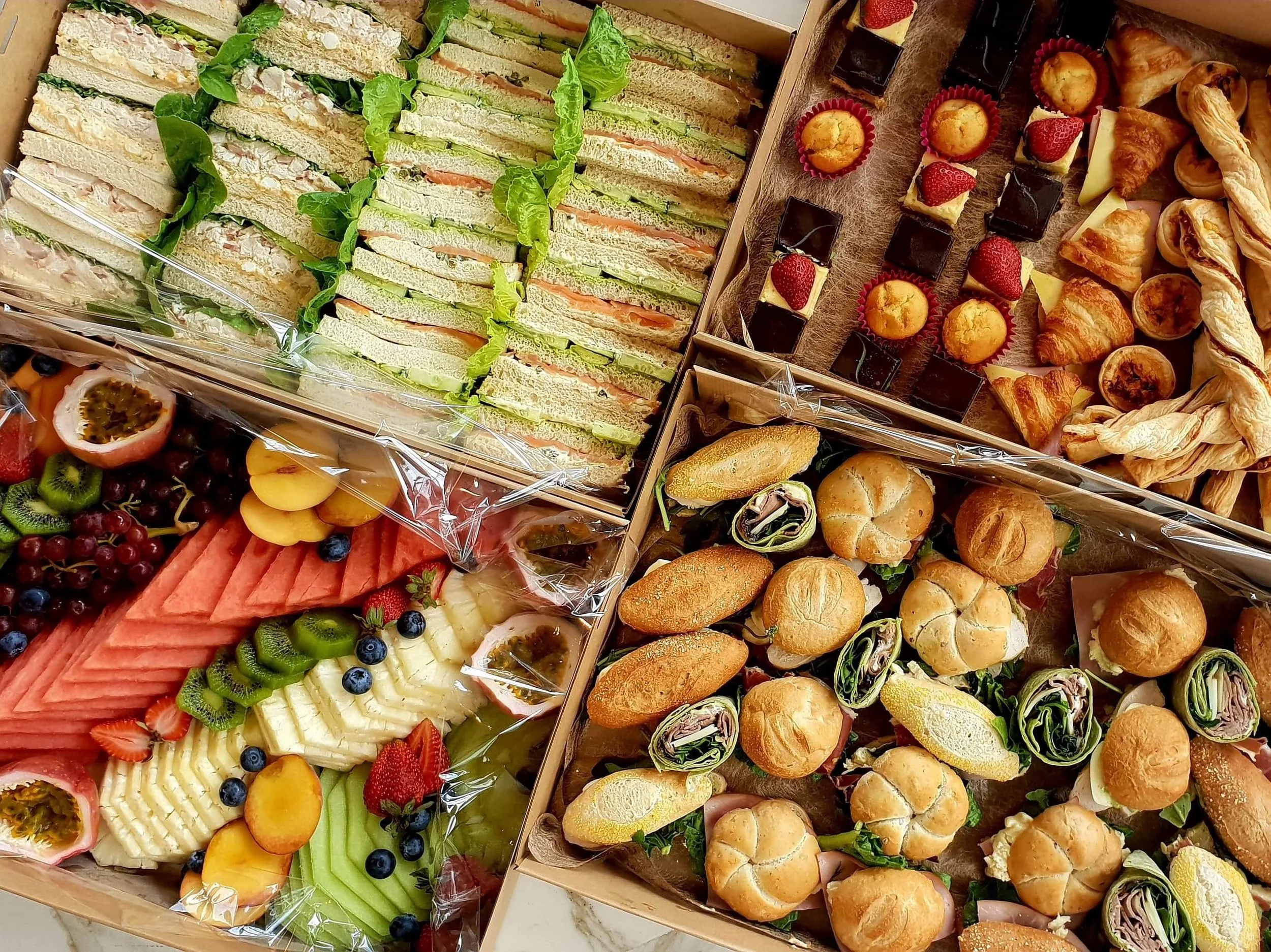 Assorted party trays with sandwiches, fruit, mini desserts, and small sandwiches.