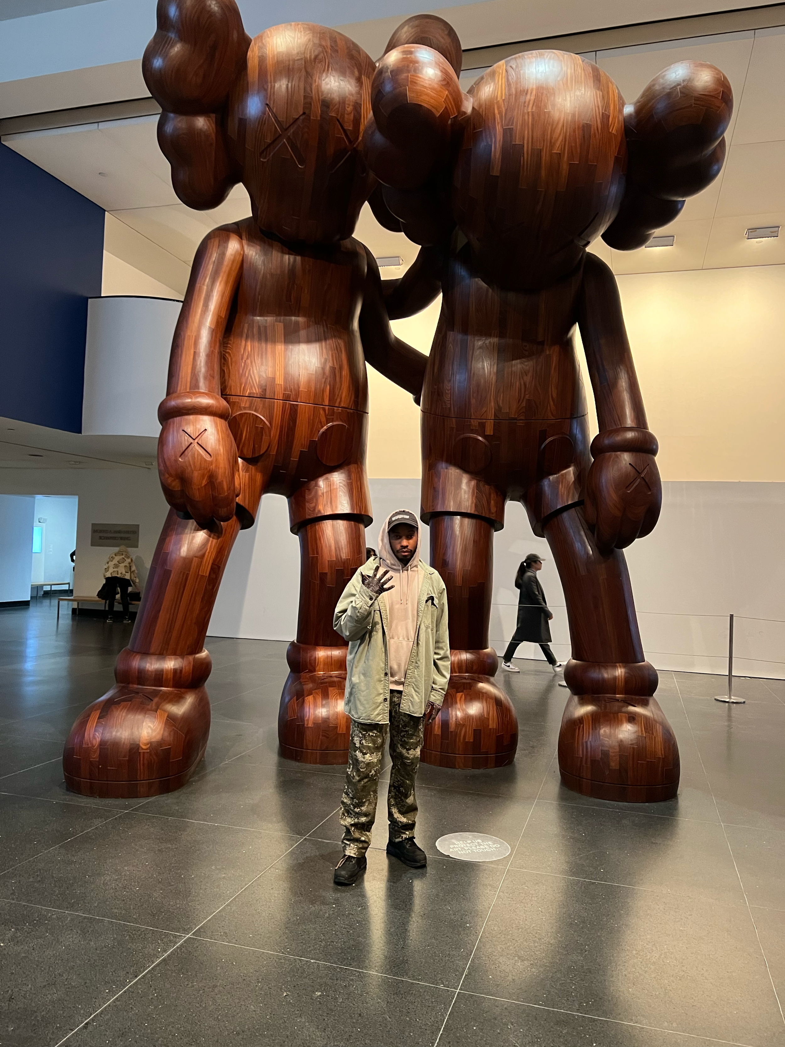Person standing in front of a large wooden sculpture of two cartoonish, muscular characters with exaggerated heads, in an indoor museum setting.