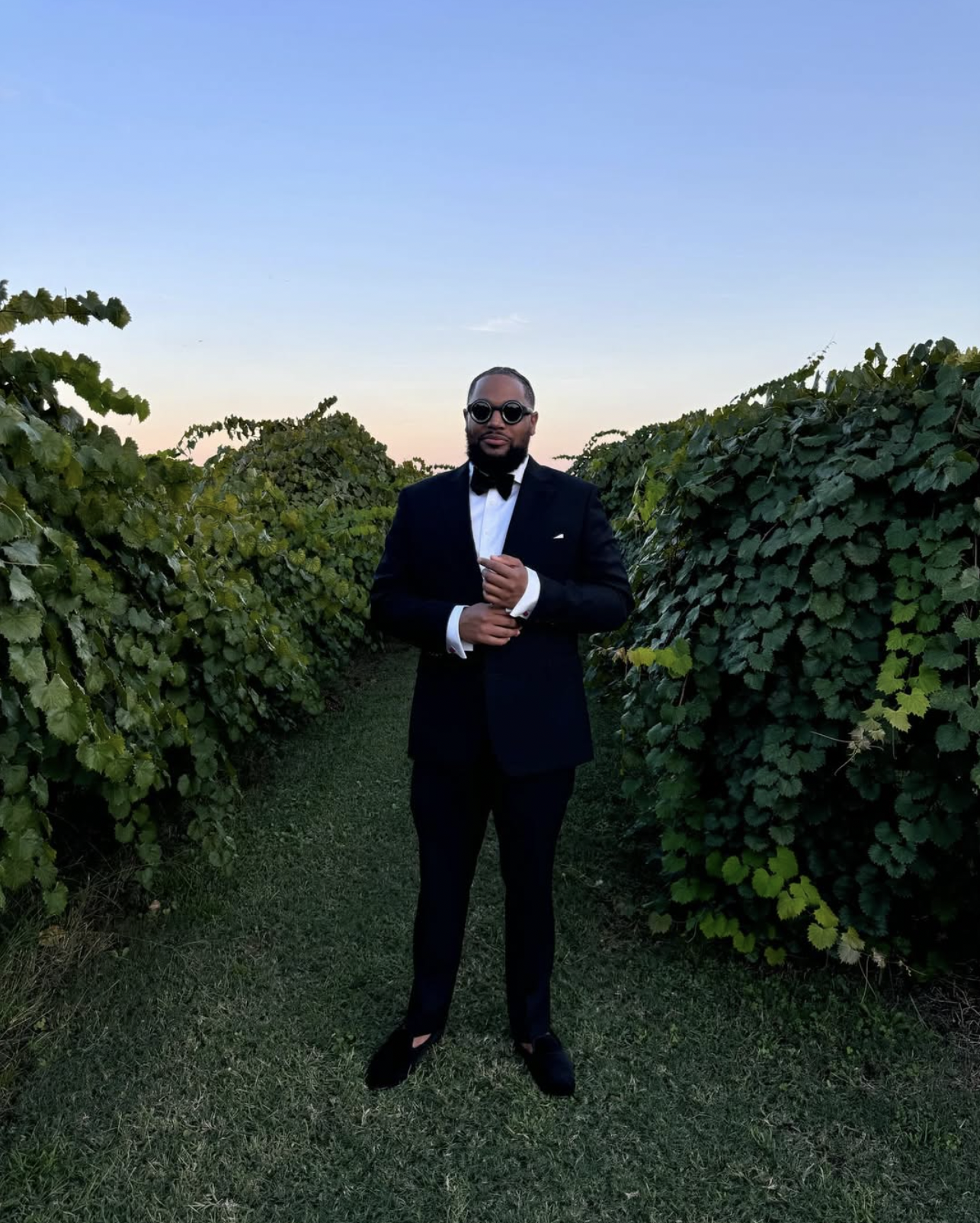 A man dressed in a black tuxedo with a white shirt, black bow tie, and round sunglasses standing in a vineyard at dusk.