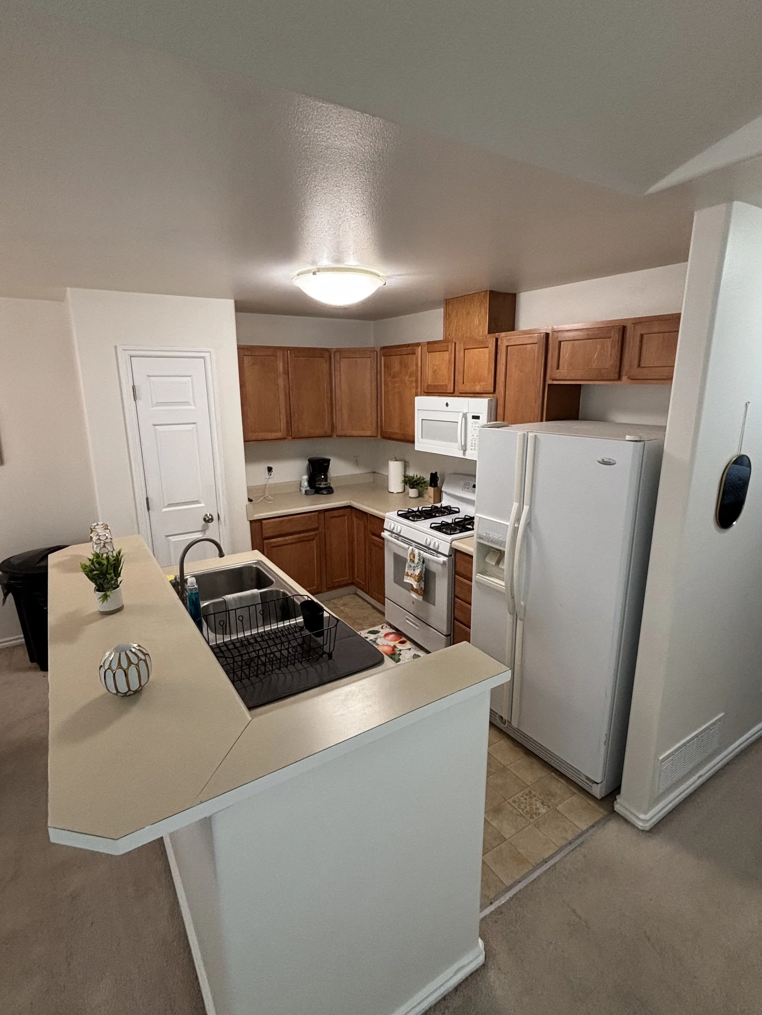 Kitchen with wooden cabinets, white appliances including a refrigerator, microwave, and stove, beige countertops, a sink with a dish rack, and a small counter with decorative items. Carpeted flooring outside the kitchen area.