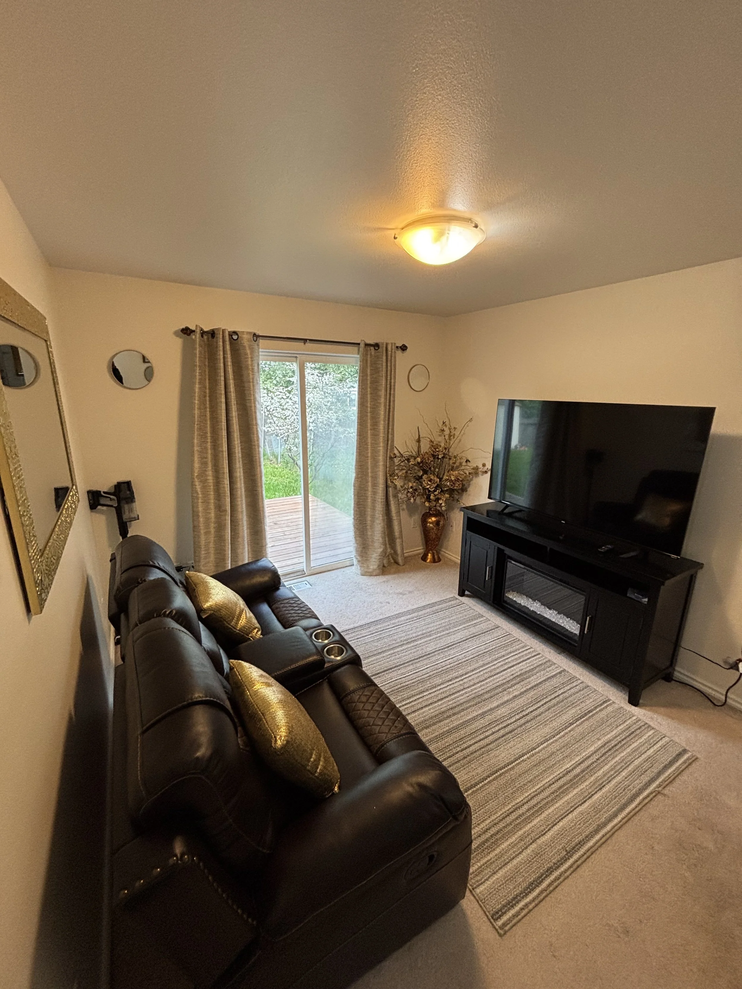 Living room with a black leather sofa, gold pillows, a striped rug, a large flat-screen TV on a black stand, a vase with dried flowers, and a sliding glass door with beige curtains leading to a patio.