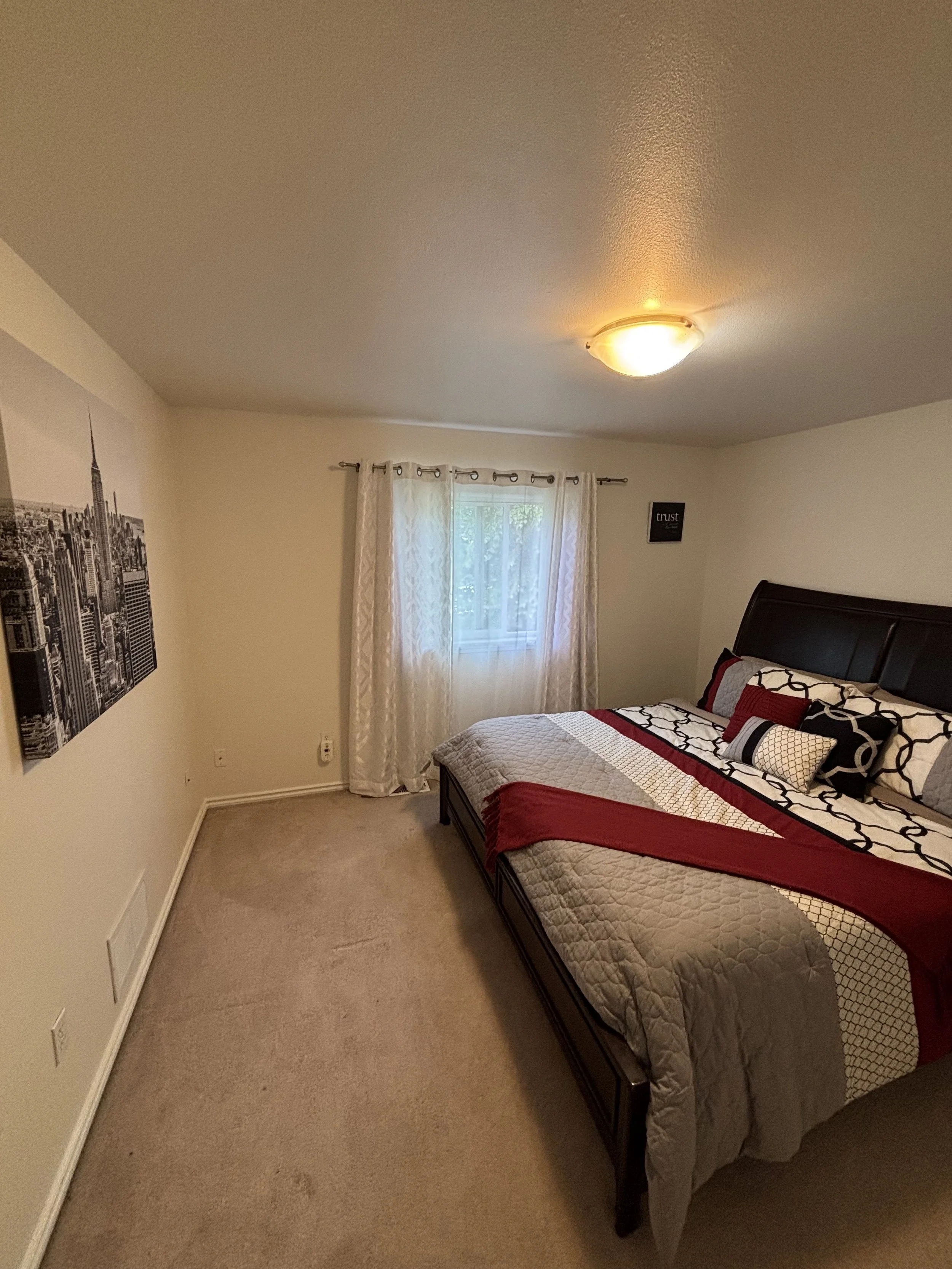 A bedroom with a bed featuring black and red bedding, a window with sheer white curtains, a black and white cityscape wall art, and a ceiling light fixture.