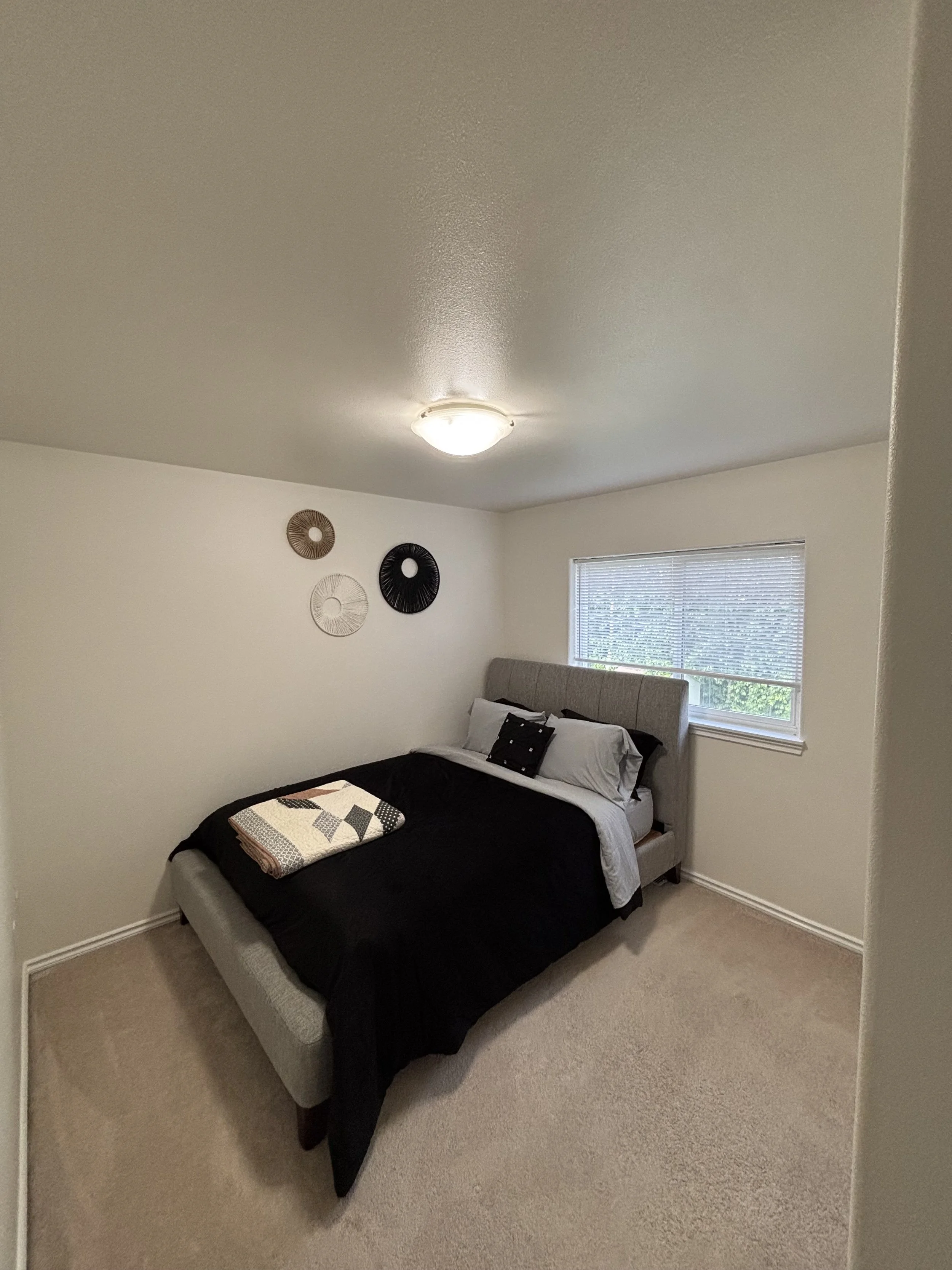 A bedroom with a bed featuring a beige upholstered headboard, black and gray bedding, and a folded quilt at the foot. There are three decorative wall hangings in circular designs on the white wall, a window with blinds, and a ceiling light fixture.