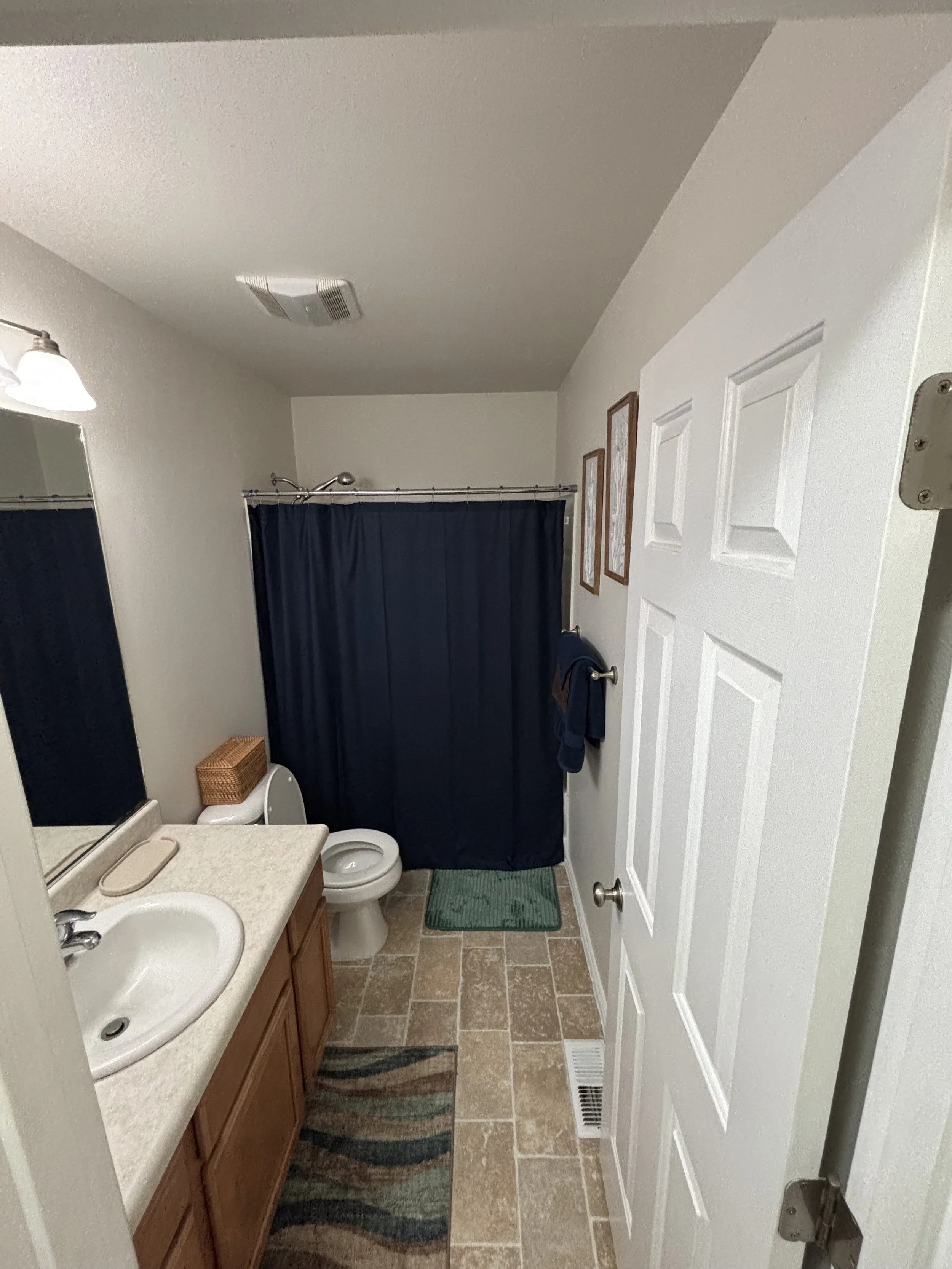 Bathroom with a beige tiled floor, a white vanity with a sink, a mirror, a toilet, a dark blue shower curtain, and framed artwork on the wall.