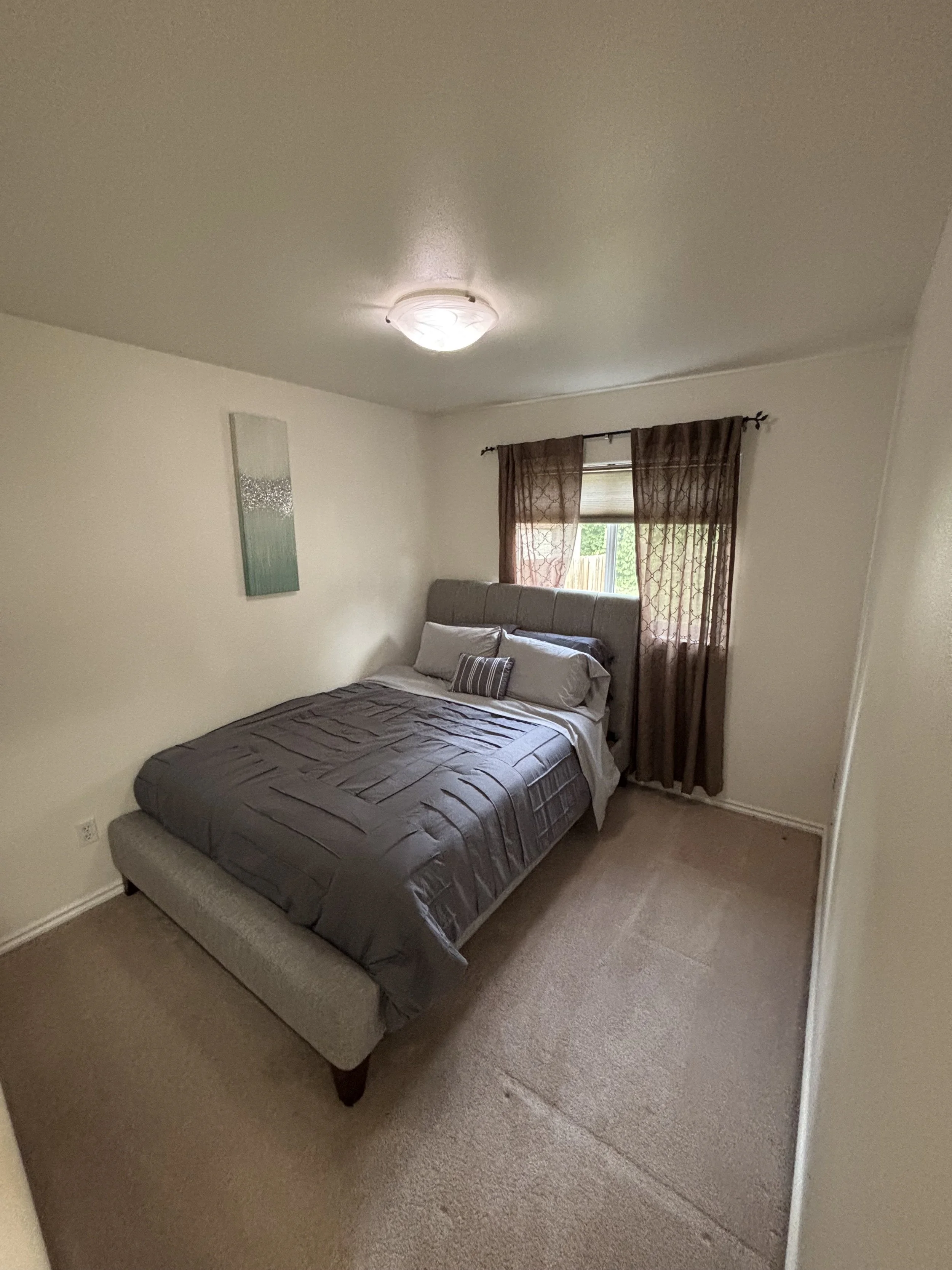 A small bedroom with a bed, window with brown curtains, beige carpeting, and a ceiling light.