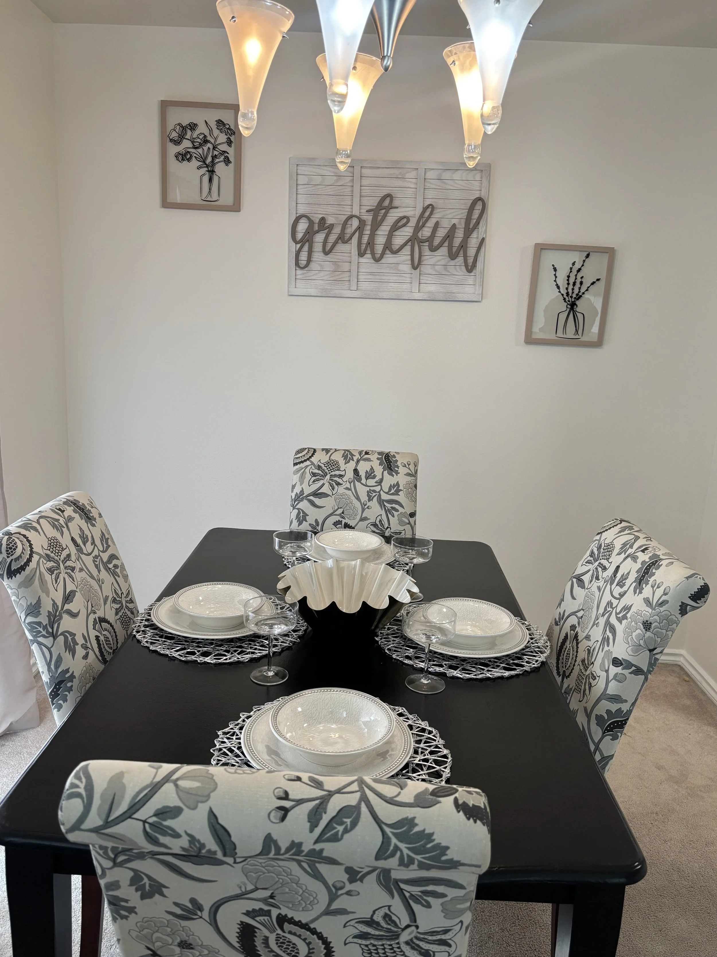 A dining room table set for four with decorative tableware, surrounded by floral patterned chairs, and a wall with framed art and a wooden sign that says "grateful."