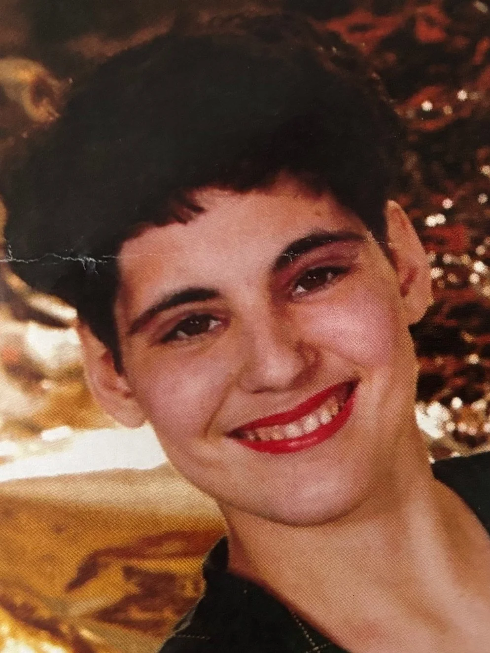 Close-up photo of a woman with short dark hair, red lipstick, and a bright smile. Background appears to have a shiny, metallic gold and copper decor.
