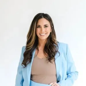 Portrait of a woman with long brown hair smiling, wearing a light blue blazer and beige top against a white background.