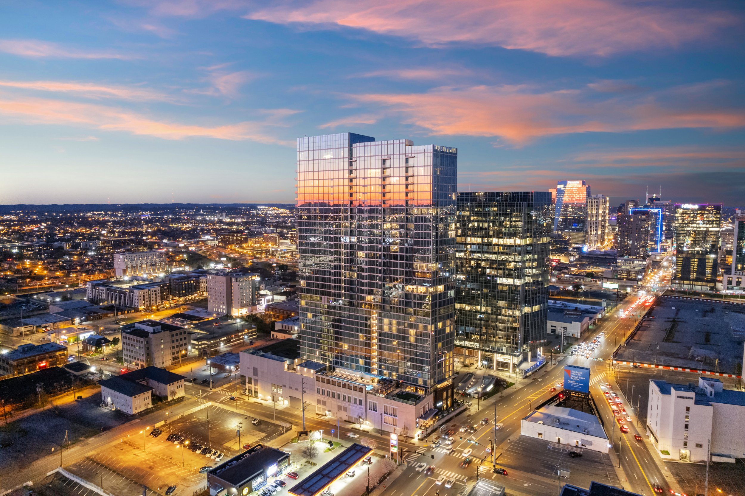 A cityscape at sunset featuring tall glass buildings reflecting the colorful sky, with busy streets filled with cars and illuminated signs.