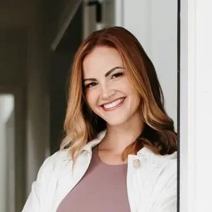 Smiling woman with long, reddish-brown hair wearing a light-colored jacket, standing indoors by a window.