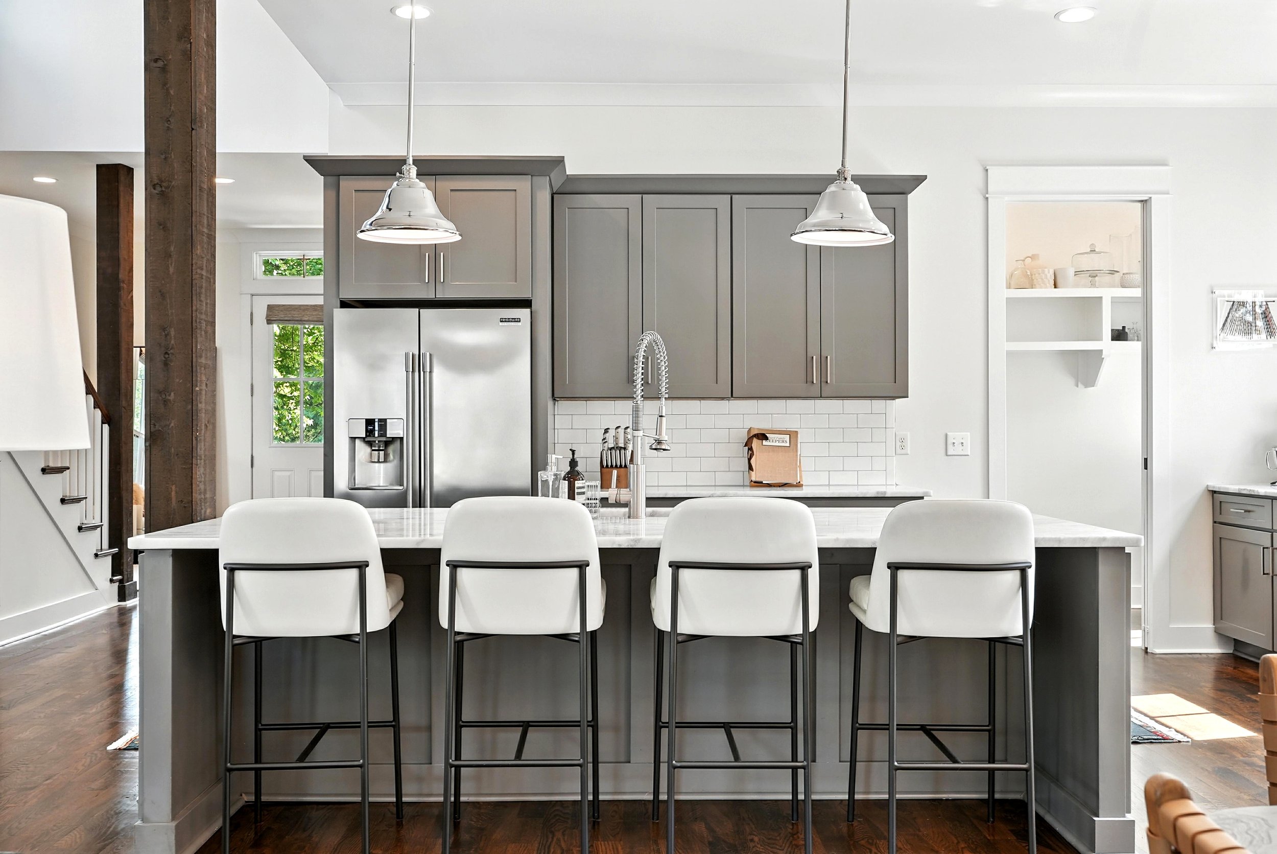 Modern kitchen with gray cabinets, stainless steel refrigerator, white subway tile backsplash, four white bar stools at a kitchen island, pendant lights, and hardwood floors.