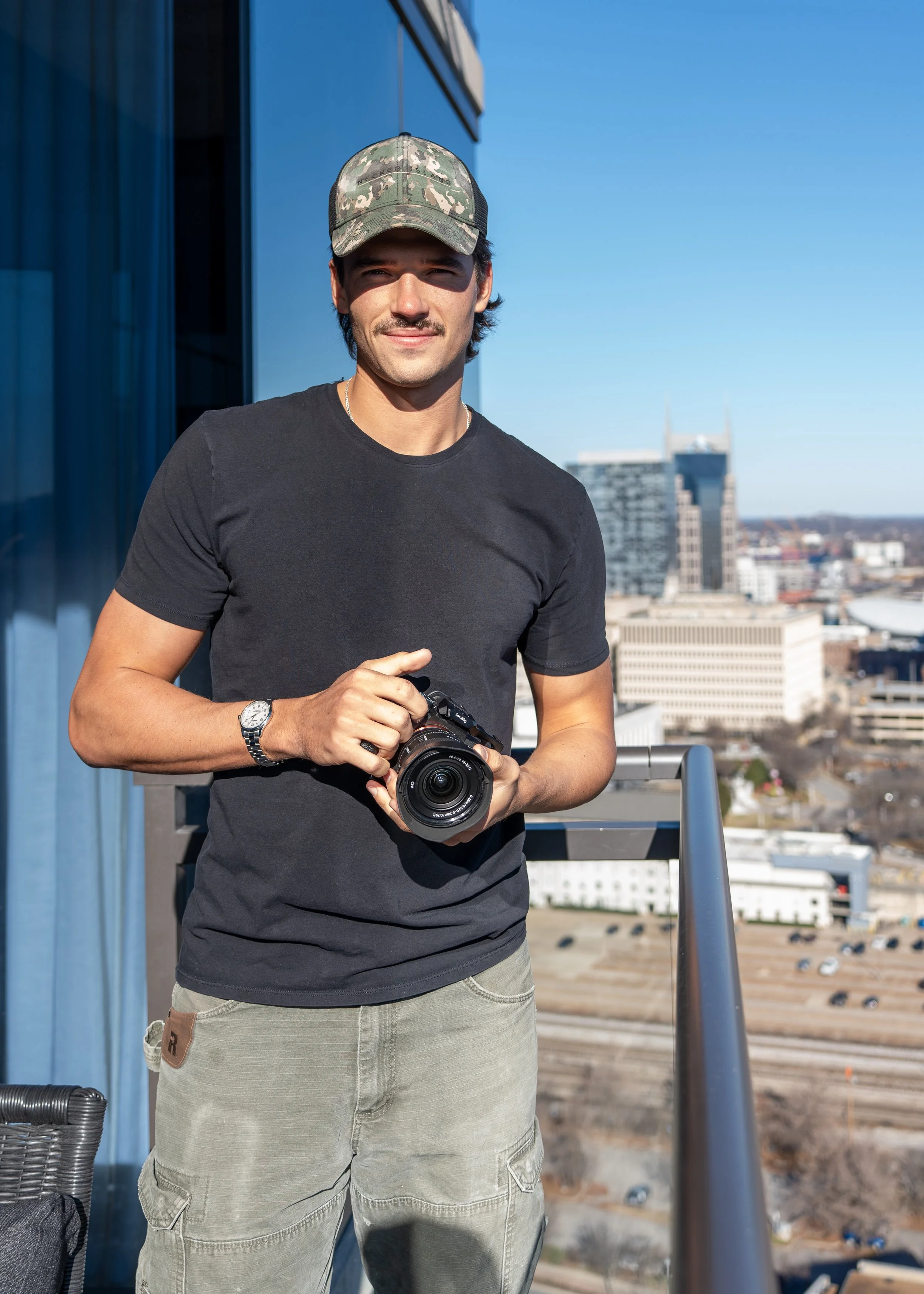 A young man standing on a rooftop balcony holding a camera, with a city skyline in the background under a clear blue sky.
