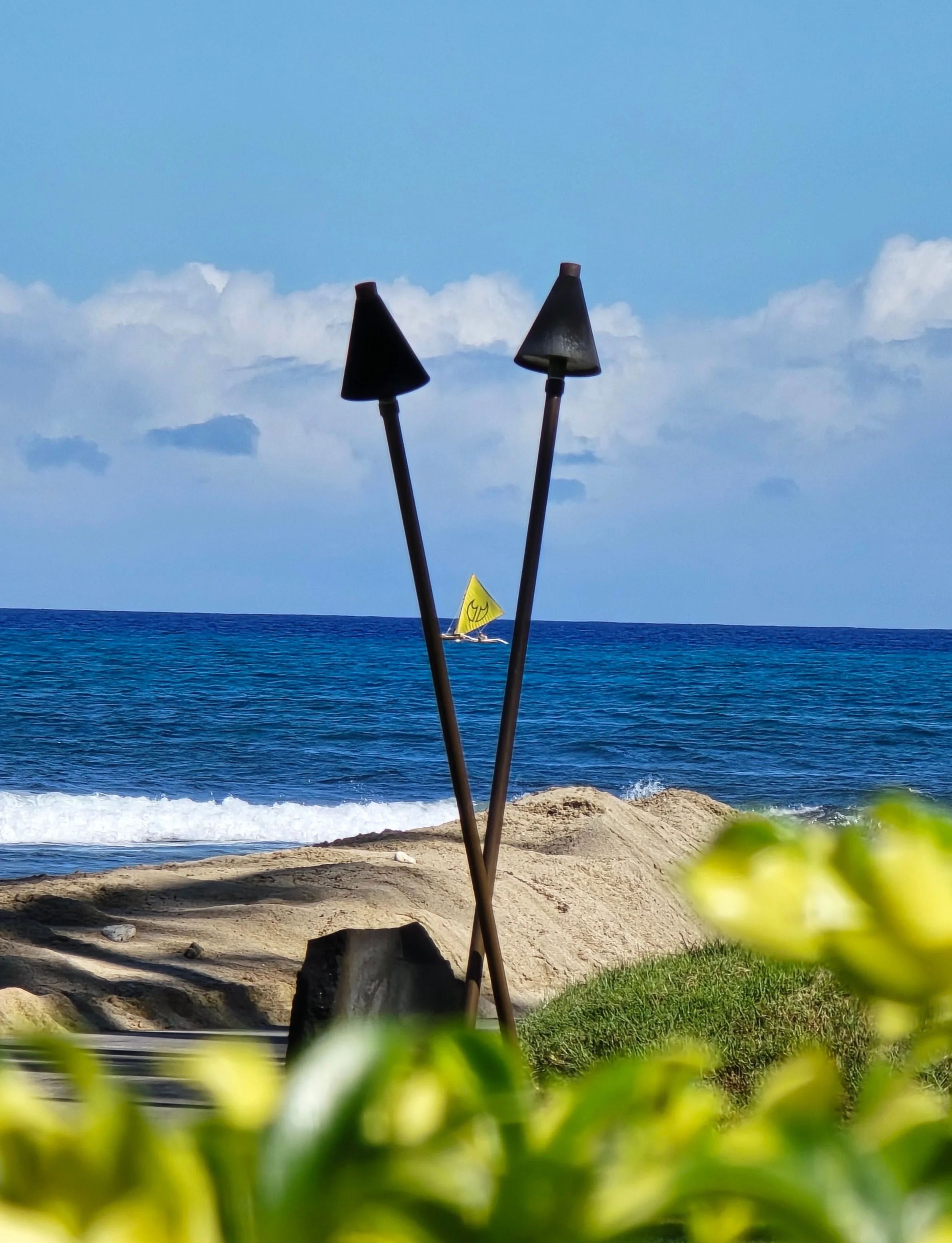 Two crossed torch holders on a grassy area near a rocky beach, with the ocean, a small sailboat with a yellow sail, and a cloudy blue sky in the background.